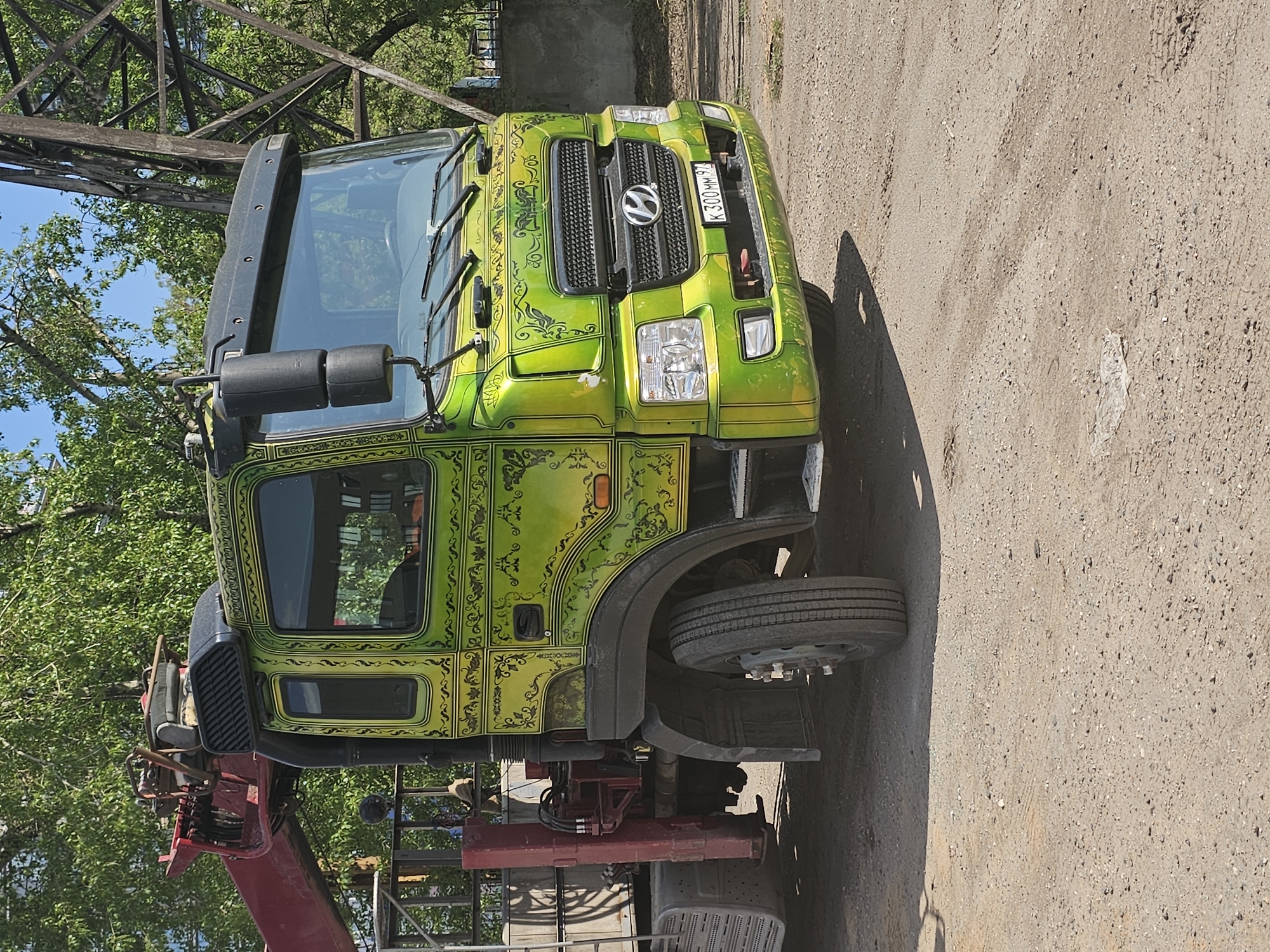 A lime green Hyundai HD170 crane truck with custom black floral and leaf ornamentation is parked on a light brown asphalt or gravel surface. The vehicle’s license plate reads "K 300 MM 97". The crane arm, painted red, is visible to the left of the truck cab. Behind the truck and to the left are green trees and a metal structure. The image was taken during the day. Based on the license plate, this photo was taken in the city of Khimki, Russia.