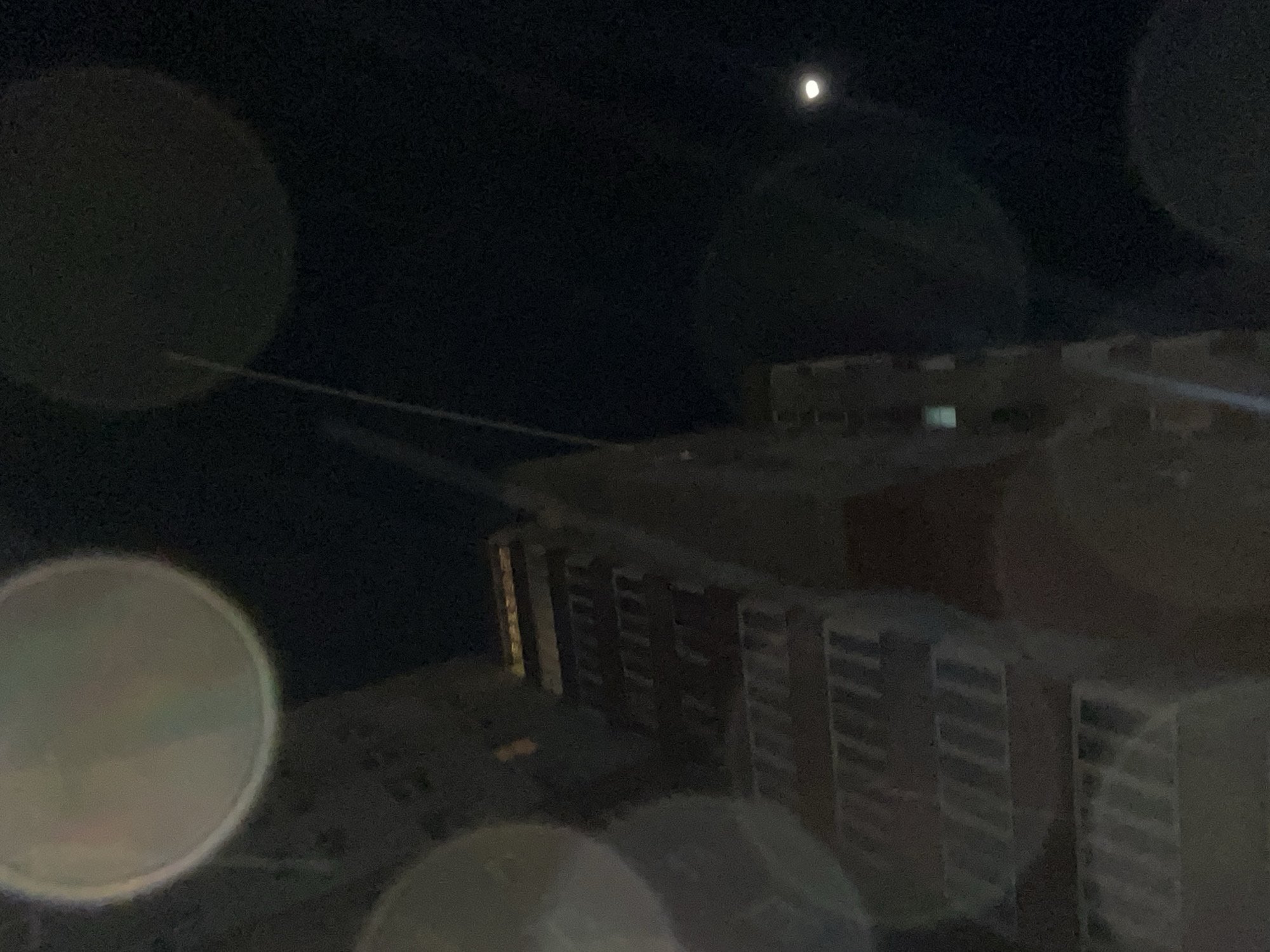 The image shows a night scene in what appears to be a city, possibly Krasnoyarsk, Russia, due to the description. The foreground is blurred with several large circular blemishes, likely due to the camera lens or environmental conditions such as water droplets. In the background, a building is visible, seemingly made of brick or a similar material, with multiple rows of windows. Above the building, a small, bright object is visible, presumably the moon, in a dark sky. A thin line crosses the frame connecting the building to something out of the frame. The overall lighting is dim.