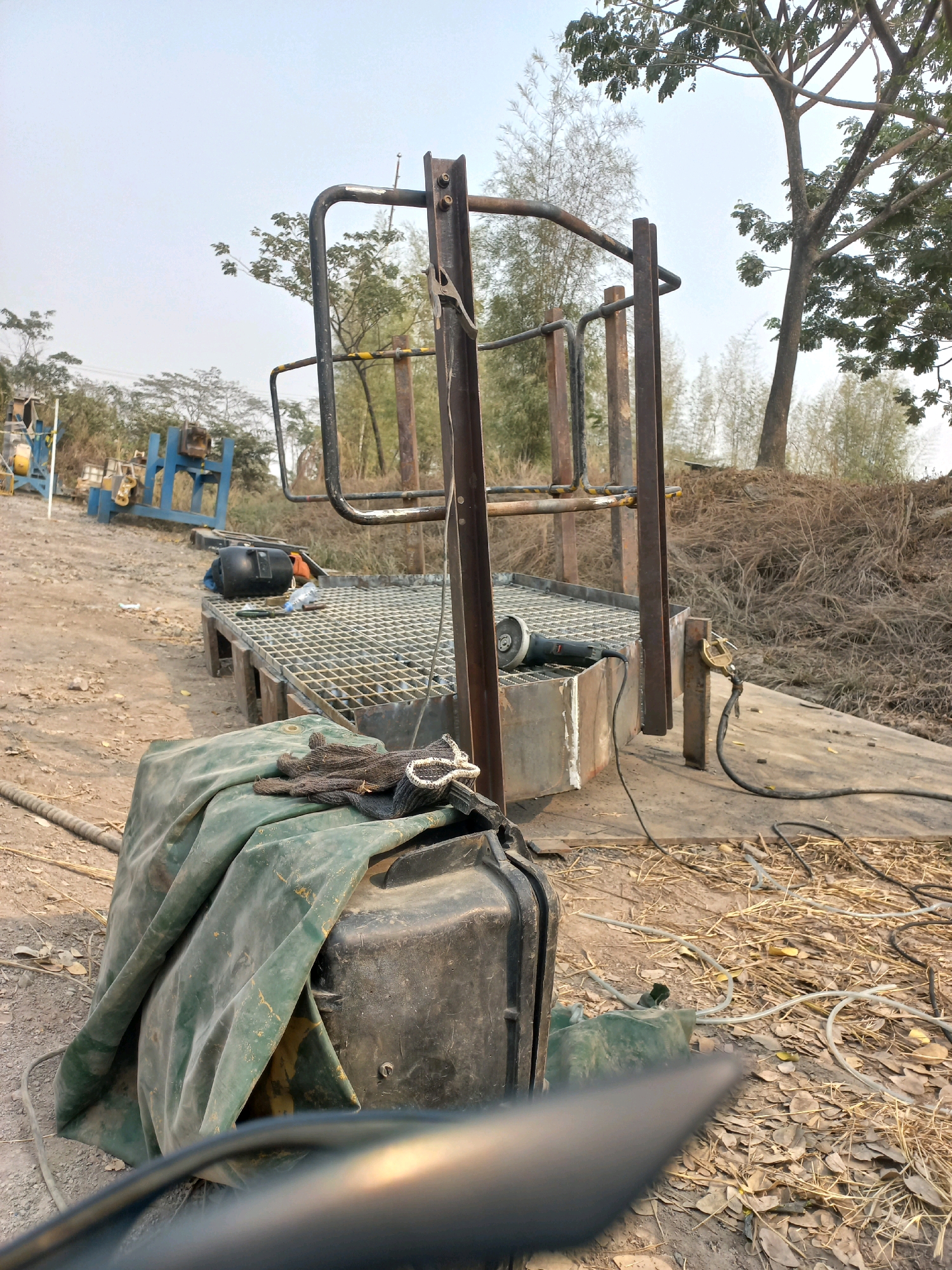The image depicts an outdoor scene, likely a construction or workshop area, in Paringin, Indonesia. In the foreground, a large black plastic container is partially covered by a green tarp, with a pair of work gloves resting on top. A thick cable snakes away from the container. Further back, a metal platform with a diamond-plate surface is supported by wooden beams. An angle grinder is visible on the platform, connected by a black cord. A metal frame structure, consisting of vertical posts and horizontal bars, stands over the platform. In the background, various pieces of industrial machinery, painted in blue and yellow, are present on a dirt-covered ground. Trees and dry vegetation surround the area, suggesting a rural or semi-developed setting. The sky is overcast. There are no people visible in the image, and no discernible actions are taking place.