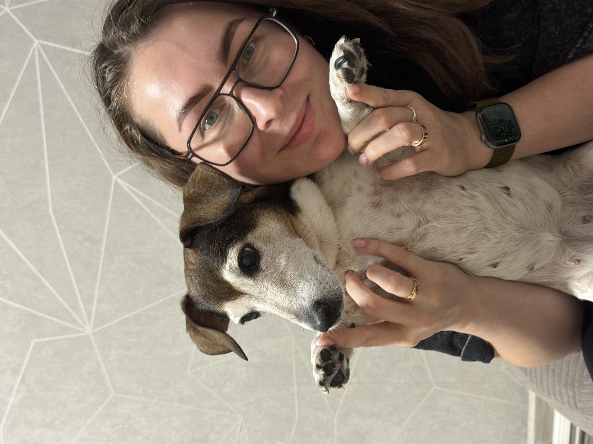 A fair-skinned female individual is depicted holding a small dog in what appears to be an indoor setting in the city of Dubovaya, Russia. The female, positioned on the right side of the frame, has long, light brown hair, green eyes, and wears black-framed rectangular glasses. She exhibits a slight smile and looks directly towards the camera. She is wearing a dark-colored top. Her left wrist bears a dark green fabric watch strap with a dark-faced smartwatch displaying "25" in green digital numerals. Two golden rings are visible on her left ring finger. Her fingernails show evidence of light pink polish.
The dog, centrally positioned and held by the female, is a short-haired breed, possibly a Jack Russell Terrier, with predominant white fur on its body and face, complemented by brown and black markings on its head, ears, and around its eyes. Its ears are floppy, and its eyes are dark. Its front paws, with dark pads and nails, are held by the female's hands. The dog maintains a neutral expression, gazing slightly to the viewer's left.
The background consists of a light-colored wall (off-white or light gray) featuring a repeating geometric pattern of thin, white lines forming polygons. The lighting in the scene is consistent.