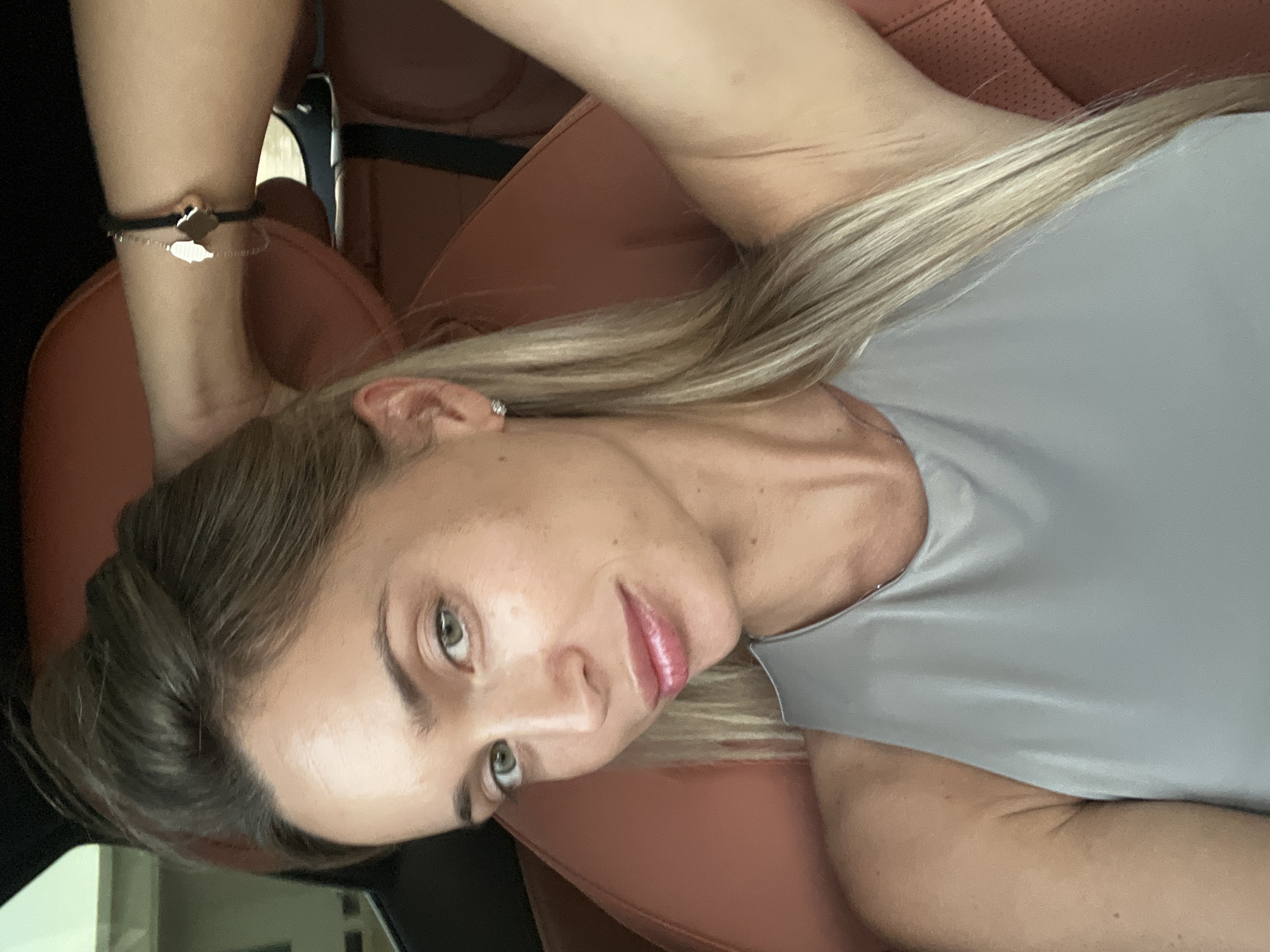 The image shows a woman with blonde hair and green eyes sitting in a car. She is wearing a grey sleeveless top and a bracelet on her left wrist. Her hair is pulled back, and she is looking at the camera. The car's interior is visible, with brown leather seats. The image appears to be a selfie taken in the car. The location is not specified in the image or the provided context.