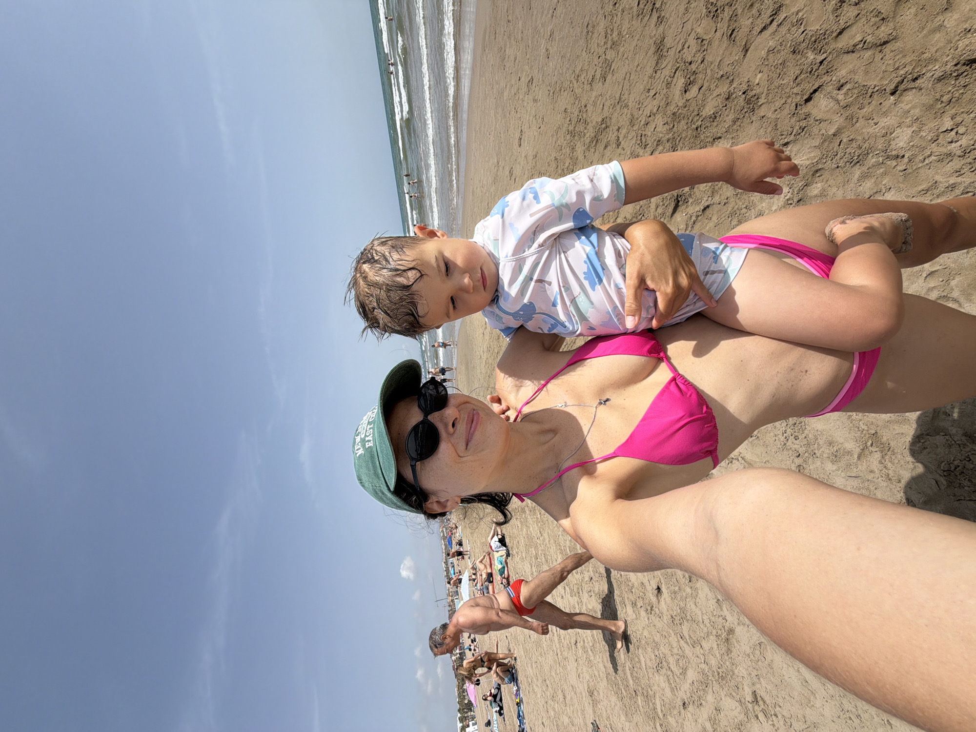 In a first-person, eye-level, wide-angle shot, a woman in a pink bikini and a green baseball cap takes a selfie on a sandy beach with a young child she is holding. The woman has dark hair, sunglasses, and is smiling. The child, wearing a light blue and white rash guard with dinosaur patterns, looks down with a neutral expression. In the background, other beachgoers are visible, some sunbathing, others walking or standing near the ocean. The sea is calm with small waves, and the sky is clear with a few wispy clouds. The sand is light brown and appears dry. The scene suggests a sunny day at the beach.