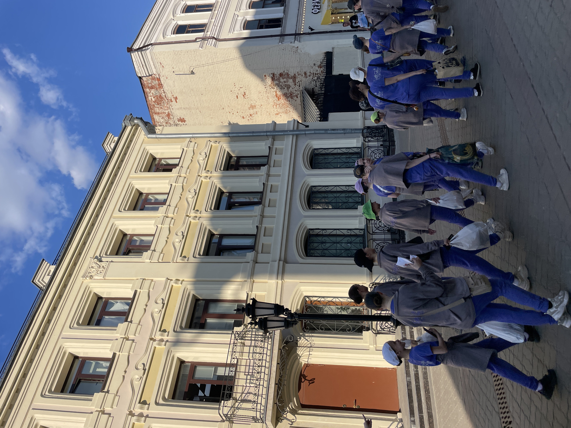 A group of people are walking on a sidewalk in front of buildings. The buildings are located in Kazan, Russia, in the historic center. The buildings are a light yellow color with ornate architectural details. The people are wearing blue pants and gray tops. They are carrying bags and papers. They are walking in a line. The sky is blue and there are some clouds.