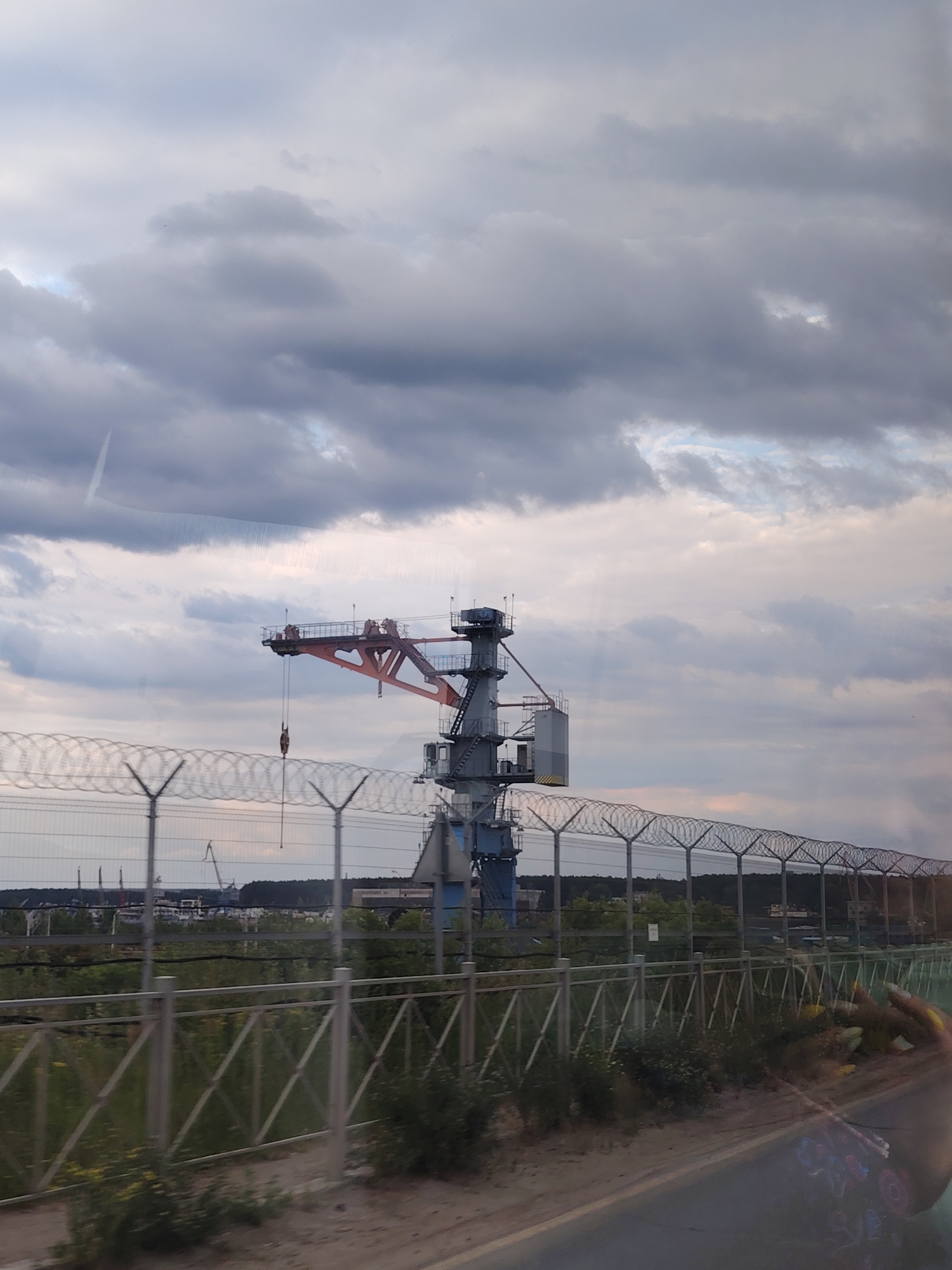 This image captures a wide shot of an industrial scene under a cloudy sky. The primary subject is a large, orange and blue crane with a visible hook hanging from its arm. The crane is situated behind a fence with barbed wire, suggesting a restricted area. In the background, other industrial structures and a treeline are visible, hinting at a port or factory environment. The sky is filled with dramatic, dark clouds, indicating overcast weather and potentially an approaching storm. The lighting suggests it is late afternoon or early evening. The presence of the fence and the imposing industrial equipment creates a sense of a functional, possibly busy, but somewhat stark environment. There is no visible text in the image.