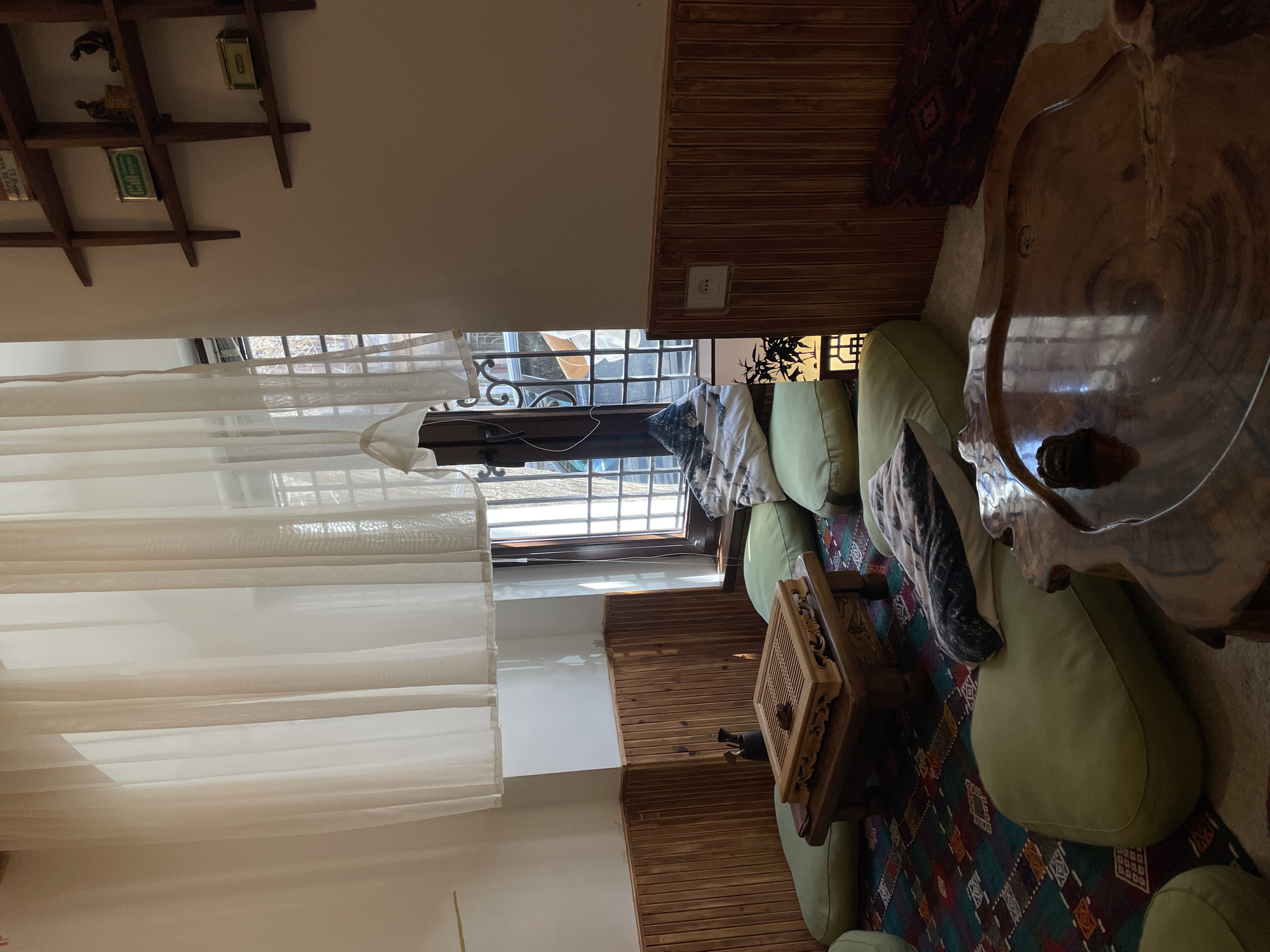 The image depicts an inviting indoor room, likely a lounge or living area, characterized by its low-profile seating and rich wooden elements. The perspective is slightly elevated, looking across the space.
In the foreground and to the right, a large, irregularly shaped coffee table made from a thick, polished wooden slab, possibly a tree trunk cross-section, serves as a prominent centerpiece. Light green floor cushions or low-slung sofas are arranged around this table and extend towards the left, providing ample seating. On the floor beneath these cushions is a colorful rug adorned with geometric patterns in shades of blue, green, red, and white. A smaller, square wooden table with intricate carvings and a lattice top is placed between some of the green cushions, holding a dark, small object.
The walls feature horizontal wooden slat wainscoting along the lower half, transitioning to plain light-colored paint on the upper sections. A wide window runs along the left side of the room, allowing bright natural light to flood the space, indicating daytime. White sheer curtains partially cover the window, and through them, a decorative metal security grill is visible. Beyond the grill, an outdoor scene unfolds, revealing some greenery, parts of a building, and the lower half of a person (wearing jeans) walking or standing outside.
On the back wall, to the left, a set of rustic wooden shelves are mounted, holding several small items. Among these are two rectangular tins: one green with what appears to be Arabic or Farsi script, possibly "شاه پسند" (Shah Pasand), and another gold-colored one marked "NAJAD" or "NAJD," likely brand names. A white electrical outlet is visible on the wooden wainscoting near the window. The room conveys a cozy, comfortable, and traditional ambiance. No specific activity is depicted within the room itself.