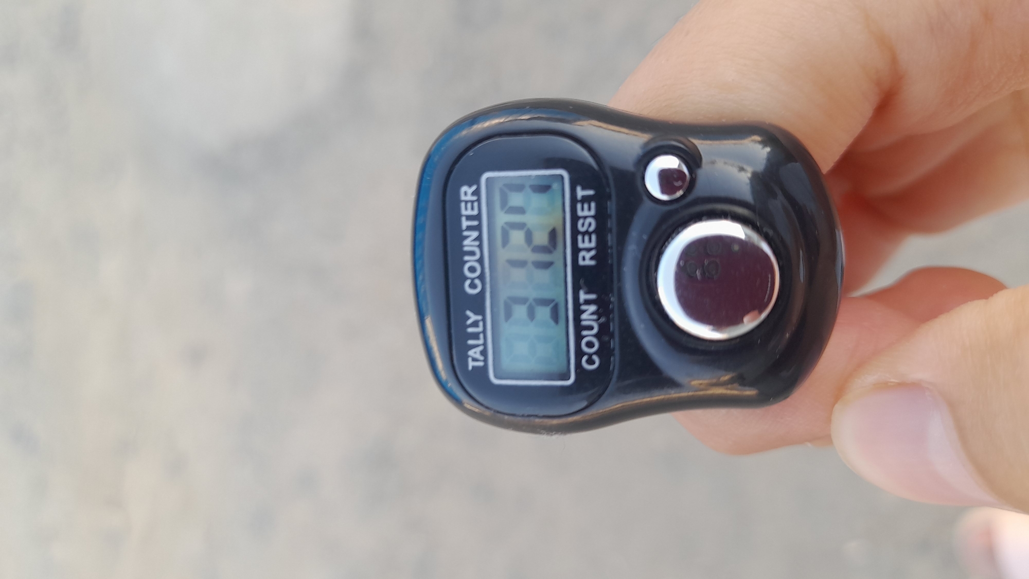 A tally counter is held in a person's hand against a blurry, light-colored background that appears to be a paved surface. The tally counter is black with a digital display showing the number 9372. Below the display are the words "COUNT" and "RESET". A large silver button and a smaller silver button are visible on the face of the tally counter. The image is taken from a close-up perspective, focusing on the tally counter and the hand holding it. The background is out of focus, providing no discernible details about the location or environment.