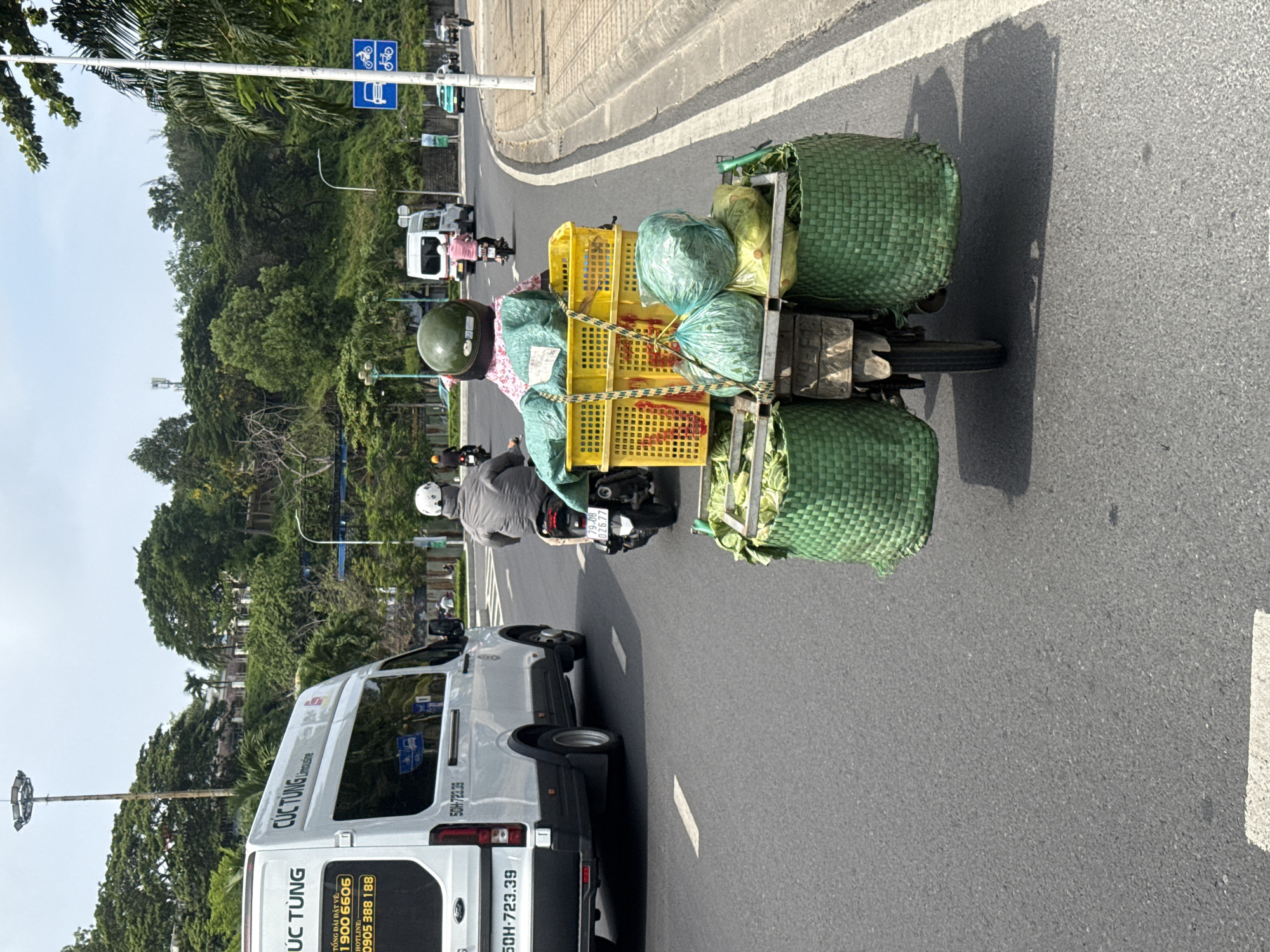 In Bac Nha Trang Ward, Vietnam, a three-wheeled motorcycle is traveling on an asphalt road, laden with agricultural produce. The motorcycle is equipped with two large, green woven baskets on either side, filled with leafy vegetables. On top of these baskets are two yellow plastic crates, also containing vegetables, with some wrapped in green plastic bags. A person, partially visible, is seated on the motorcycle, facing away from the camera. A white van with "CÚC TUNG Limousine" branding is in the left lane of the road, also moving in the same direction. The background features a tree-covered hillside, buildings, and streetlights. The sky is overcast.