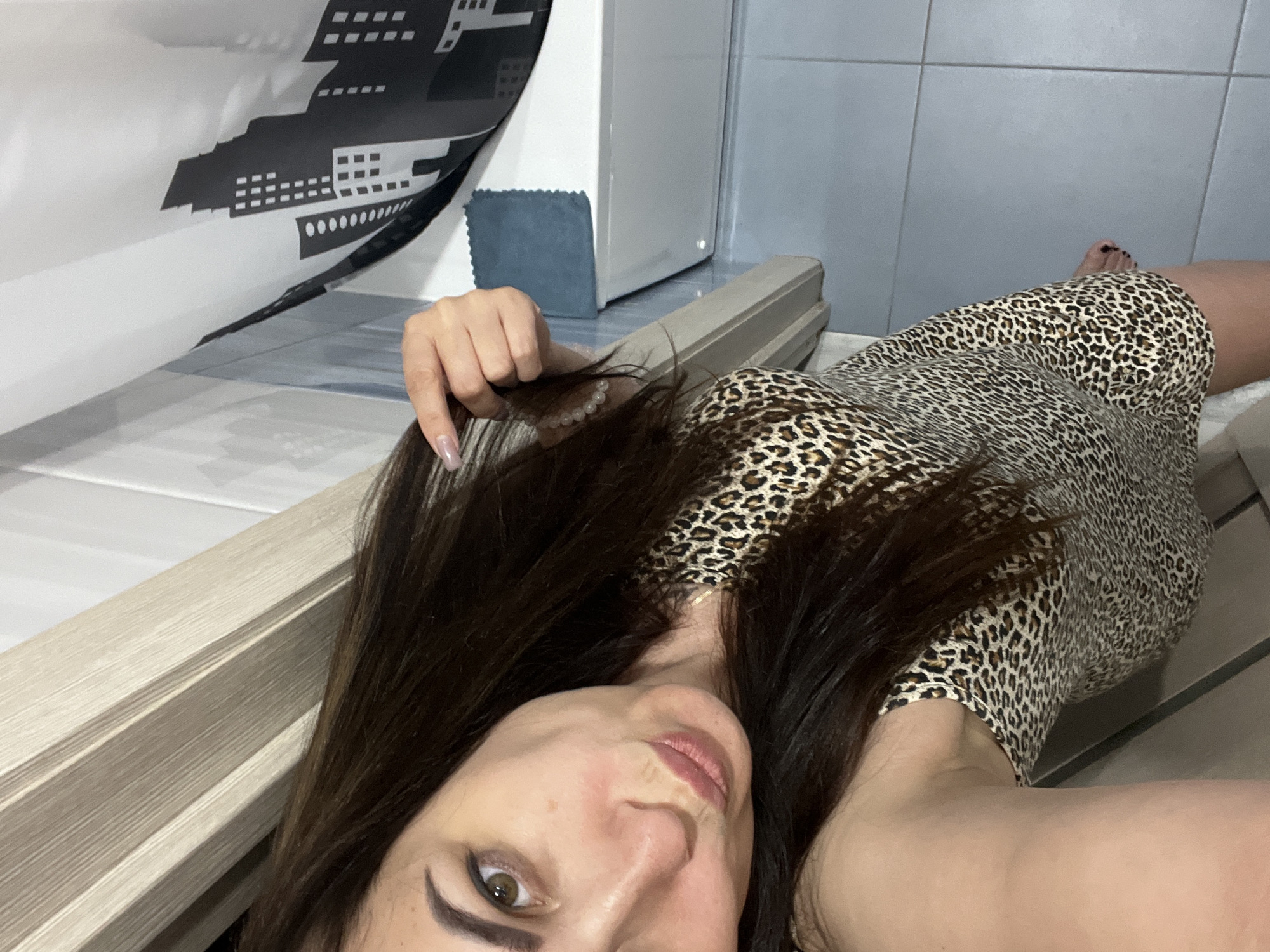 A woman is taking a selfie in a bathroom. She is wearing a leopard print dress and has long dark hair. She is pouting her lips and holding a strand of her hair. In the background, a shower curtain with a cityscape design is visible. The bathroom floor is tiled in a light blue color.