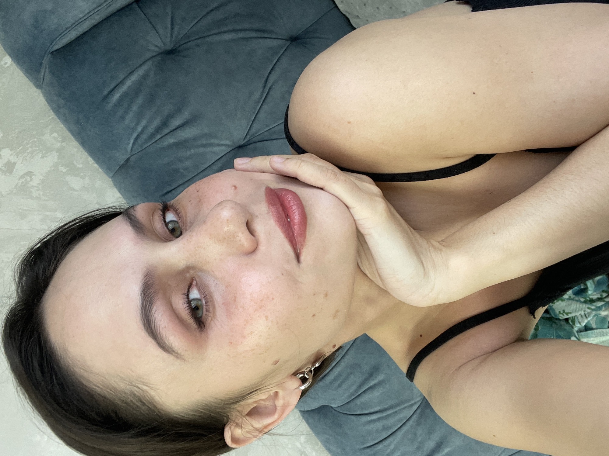 A woman is lying on a gray cushioned surface, possibly a sofa, indoors. She is wearing a black top with thin straps. Her head is tilted, and she rests her chin on her hand. She has brown hair, fair skin with freckles, and light green eyes. She is wearing light pink lipstick and has a silver earring. The background includes the gray cushioned surface and a glimpse of a patterned fabric. There is no indication of a location in Izhevsk, Russia, or any city, within the image.