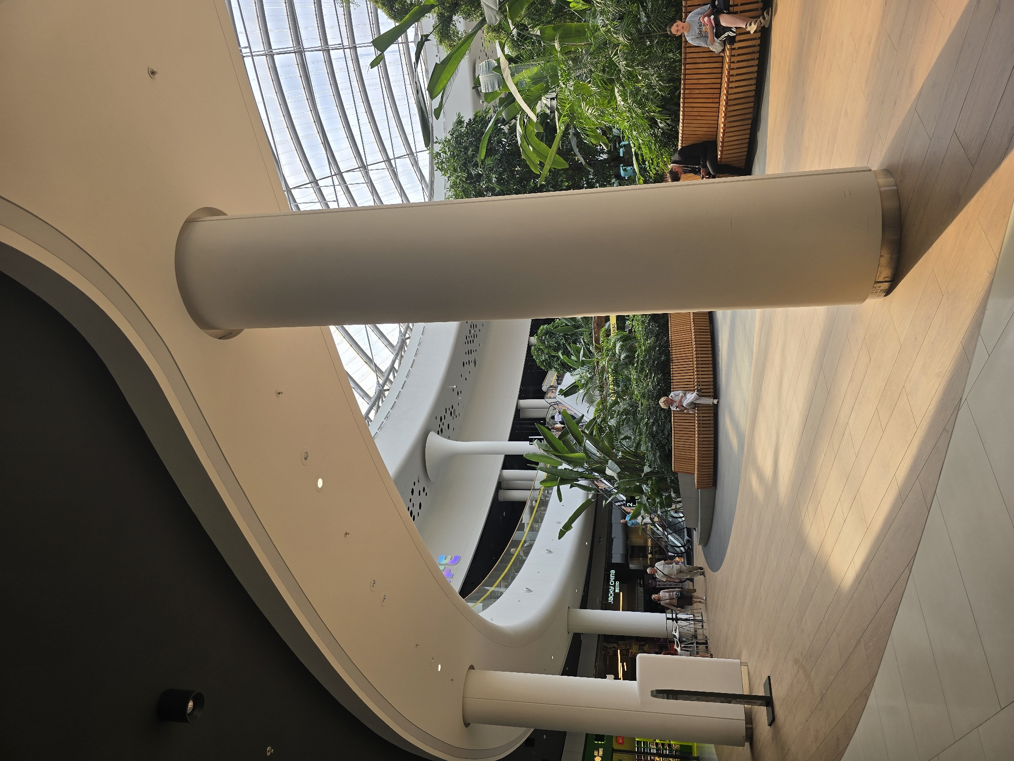 The image depicts the interior of a modern shopping mall with an atrium. The architecture features a curved white ceiling with a black section and a large, cylindrical white pillar in the foreground. The floor is made of light-colored wooden planks. In the background, there is a large green indoor garden with lush plants and trees. Several people are visible: a couple walking in the middle ground, a woman sitting on a wooden bench in the garden, and two individuals seated on a wooden bench on the right side. The overall atmosphere is bright and airy, with natural light filtering through a glass ceiling. The location is identified as a shopping mall in Kyiv, Ukraine.