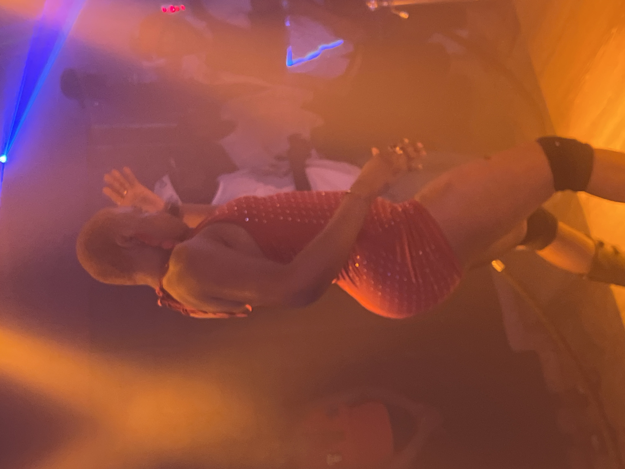 The image shows a person wearing a red dress and a black knee pad, lying on what appears to be a surface with some light and smoke in the background. The setting appears to be indoors, potentially a performance space in Howayi, Nigeria, given the prompt. The person's position and attire suggest a performance or entertainment context.
