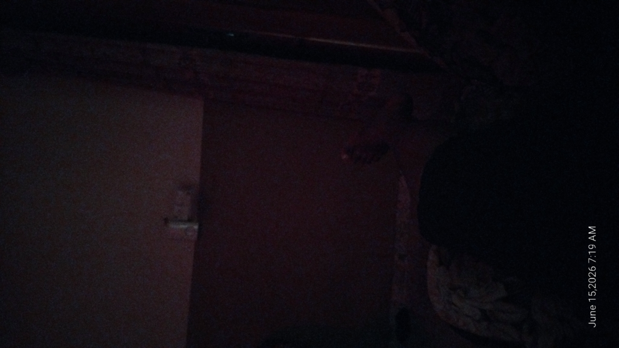 The image depicts a dimly lit bedroom interior. The focal point is a person lying down, mostly obscured by shadows, with only a portion of their body and head visible. The environment suggests a domestic setting, with what appears to be bedding and possibly a wall with some indistinct objects mounted on it in the background. The lighting conditions are poor, creating a dark and somber atmosphere. The date and time stamp "June 15, 2026 7:19 AM" is overlaid on the bottom left corner of the image. There is no information in the image to indicate the specific location within Howayi, Nigeria.