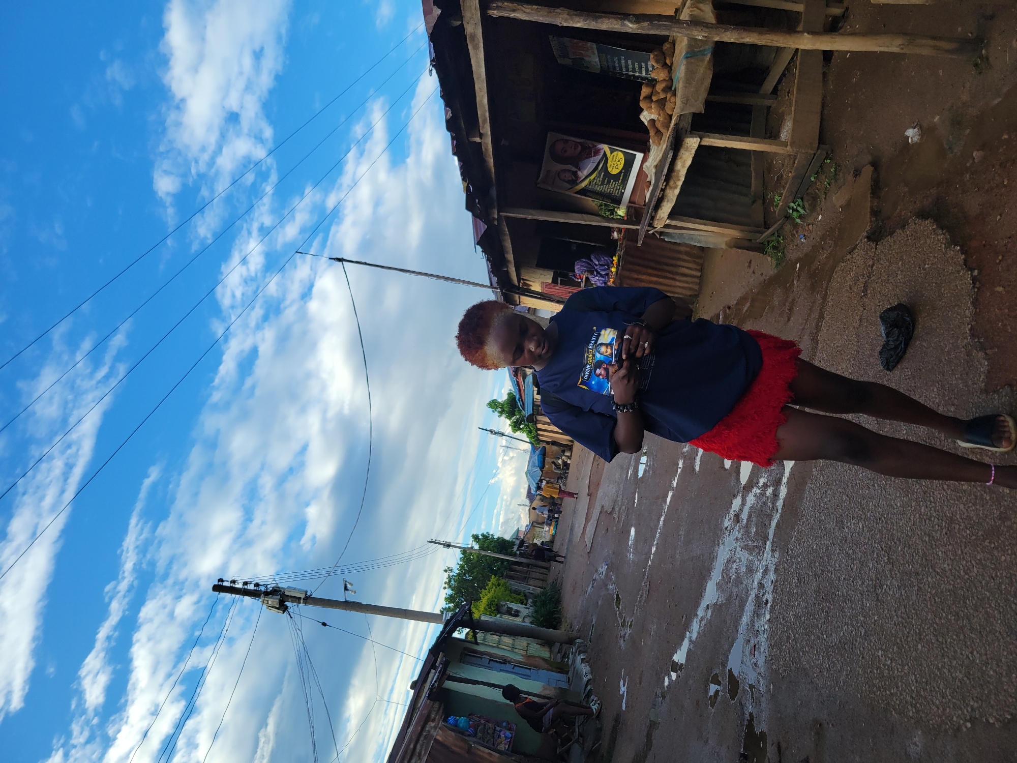In this full-shot, eye-level outdoor medium shot, a young Black woman with short, reddish-blonde hair stands on a dirt and gravel road, looking down at her phone. She is wearing a dark blue t-shirt with a graphic print on the front and a short, bright red, textured skirt. Her left arm is bent as she holds the phone, and her right arm is by her side. She wears black slides on her feet.
The environment appears to be a street in a developing area. To the left of the woman, a row of corrugated metal and wood structures, possibly market stalls or small shops, are visible. One stall has a person sitting in the doorway. The ground is wet, with puddles reflecting the cloudy blue sky. Overhead, power lines crisscross the sky, supported by a utility pole. To the right of the woman, another similar stall is present, with a table displaying a pile of root vegetables. A poster with images of women is attached to the stall. In the background, more stalls and people are visible further down the street. The lighting suggests it is daytime, possibly late afternoon, with soft light and shadows.