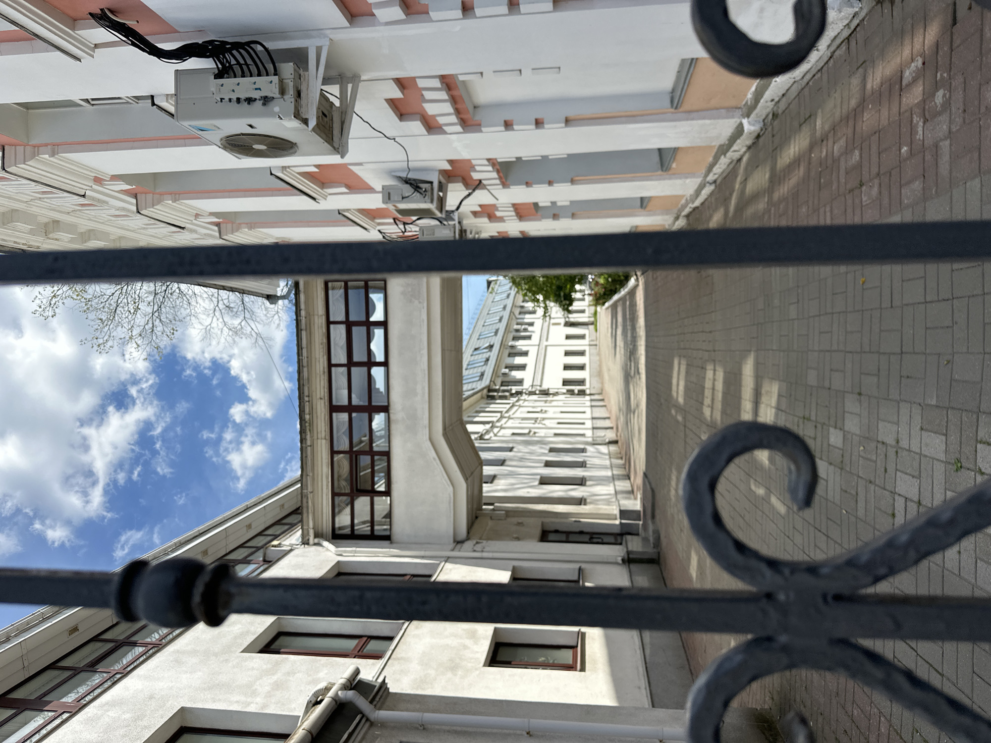 The image shows a narrow pathway between two buildings, seen through a black wrought iron fence. The buildings appear to be part of an institutional complex, possibly a school or hospital, with a skybridge connecting them on the left side of the image. The building on the right has a light pink and white facade with rows of windows and air conditioning units. The pathway is paved with brick and is illuminated by sunlight, creating distinct shadows from the buildings. The sky is blue with scattered white clouds. No people are visible in the image. The location is described as Unknown, Unknown.