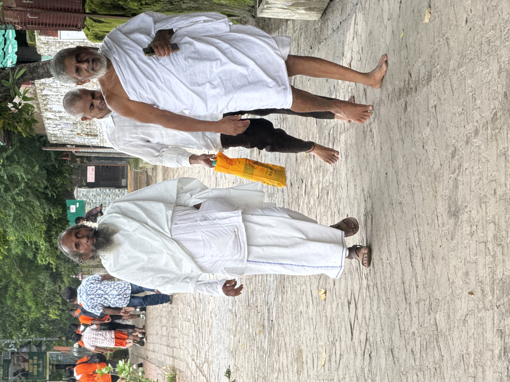 Three men are walking on a cobblestone path in Rishikesh, India. The man in the foreground, on the right, is elderly with a white beard and is wearing a white robe draped over his left shoulder and around his body. He is holding a dark-colored object, possibly a phone, in his right hand. To his left, another man, also elderly, is walking and looking downwards. He is wearing a white shirt and black pants. Further to the left, the third man, who appears younger and has a beard, is walking and looking forward. He is wearing a white robe and a white cloth wrapped around his head. He is carrying a bright orange bag in his left hand. In the background, other people are walking, some wearing orange t-shirts and others in patterned shirts and jeans. The environment includes greenery with trees and buildings lining the street. The day appears overcast.