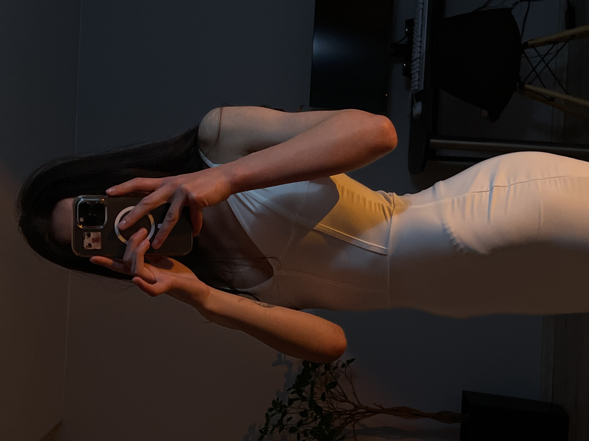 A vertical shot captures a person, likely female, in a dimly lit indoor setting, possibly in Ternopil, Ukraine. The person is wearing a white, sleeveless, form-fitting dress and is holding a smartphone with both hands, obscuring their face, as if taking a mirror selfie. Their long, dark hair cascades over their shoulders and back.
The lighting is low, with a warm light source illuminating the person's right arm, shoulder, and parts of the dress, creating highlights and shadows that emphasize the contours of their body. The smartphone, held at eye level, has a clear case with a visible magnetic ring on the back. The person's fingers are wrapped around the device.
In the blurred background, a dark wall is visible, along with indistinct shapes of furniture or equipment, including what appears to be a black screen or monitor and a chair with a metallic frame in the upper right. A decorative plant or branch with sparse leaves can be seen in the lower right portion of the frame. The overall atmosphere suggests an evening or nighttime scene due to the low ambient light. No discernible text is visible in the image.