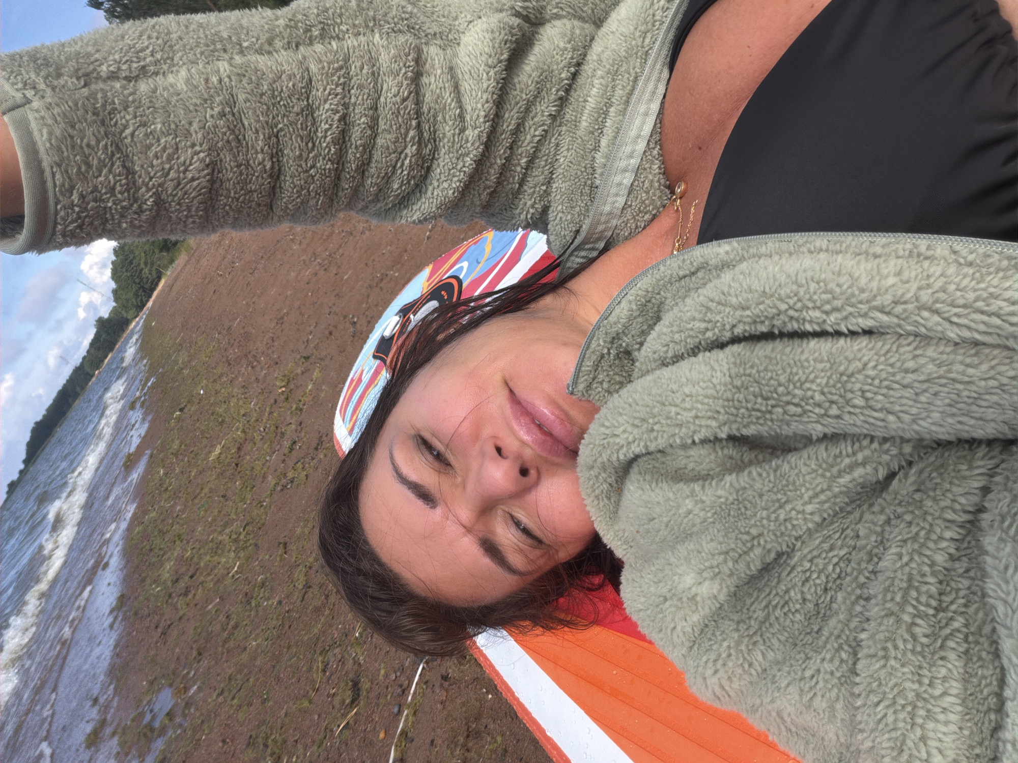 A woman lies on a colorful beach towel, covered in a fuzzy green jacket, with her left arm raised. She wears a black bikini top. The background shows a brown, sandy shoreline with waves of water lapping at it, and a line of trees in the distance under a cloudy sky. The location is identified as Vyazy, Russia.