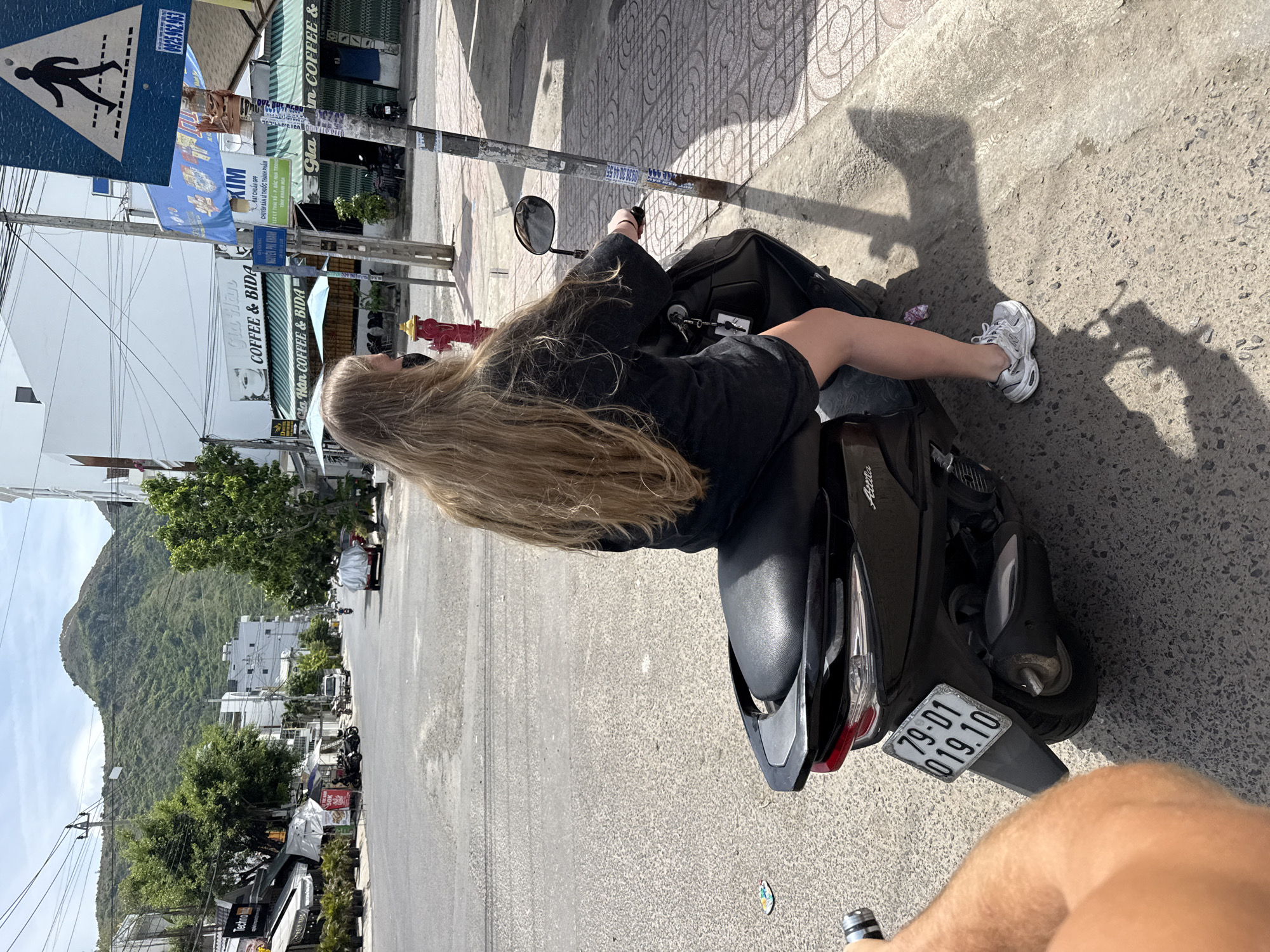 A person with long blonde hair, wearing a black t-shirt and shorts, is sitting on a black scooter on a paved street. The scooter has a license plate with the numbers "79-01 019.10". In the background, there are buildings with awnings, trees, a large green hill, and a pedestrian crossing sign. The sky is clear and blue. The scene is located in Bac Nha Trang Ward, Vietnam.