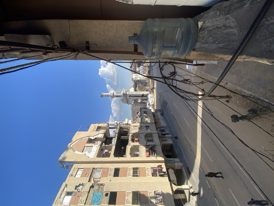 An elevated view captures a war-damaged urban street scene in Khan Yunis, Palestinian Territory, under a clear blue sky with scattered white clouds. In the immediate foreground to the right, a large, transparent blue water cooler bottle rests on a concrete ledge. Numerous black electrical wires crisscross the frame, some attached to a rusted metal railing, partially obstructing the view.
To the left, a multi-story, beige apartment building exhibits severe structural damage, with large sections of its facade missing, exposing internal rooms, rebar, and debris. A blue tarp covers an opening on one of the upper floors, while laundry hangs from balconies on lower, relatively less damaged sections.
A two-lane paved road, marked with a solid white line, runs diagonally through the midground. On the road, several individuals are visible: one person in dark clothing walks in the left foreground, another walks further down the road toward the center, and two smaller figures are in the distance. A parked motorcycle with a black crate on its rear rack is situated on the right side of the road.
In the background, at the end of the road, stands a significantly damaged mosque with its white minaret largely intact but the main structure appearing partially collapsed and surrounded by extensive rubble and debris. Other damaged buildings are visible in the distant background. The scene is illuminated by bright sunlight, casting distinct shadows.