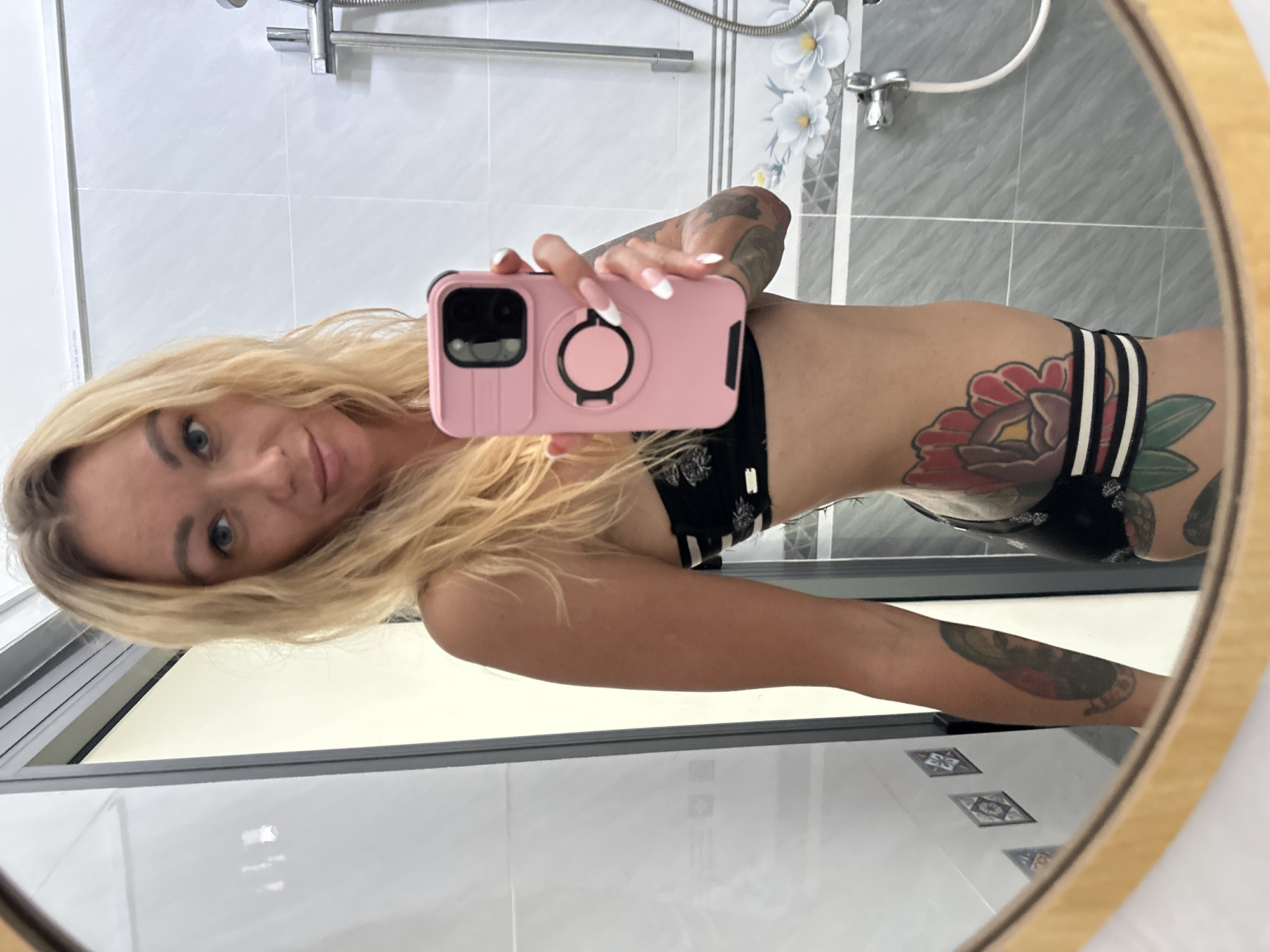 A woman with blonde hair and tattoos is taking a mirror selfie in a bathroom. She is wearing a black and white bikini. The bathroom has grey tiled walls and a white ceiling. There is a shower head visible in the background. The woman is holding a pink phone in her right hand. The reflection in the mirror shows a tiled wall with a floral pattern. The setting is likely a hotel bathroom in Mũi Né, Vietnam.