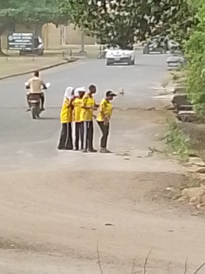 The image captures an outdoor scene on a bright, sunny day, featuring a road bordered by trees and foliage. In the midground, a group of four individuals is standing on the shoulder of the road, which appears to be unpaved or dirt, adjacent to the main paved section. All four are wearing bright yellow short-sleeved shirts.
From left to right within the group:
1. The first person wears a white head covering, possibly a hijab, and dark, loose-fitting trousers or a long skirt.
2. The second person is also wearing a yellow shirt and dark trousers, with a white head covering, possibly a cap or hood.
3. The third person wears a yellow shirt and dark trousers.
4. The fourth person, positioned slightly to the right, wears a dark baseball cap, a yellow shirt, and dark trousers. They are slightly turned, seemingly looking towards the right or down.
Behind this group, on the paved section of the road, a person is riding a motorcycle away from the viewer, heading towards the left side of the frame. The motorcyclist is wearing a light-colored, possibly beige or white, shirt.
In the background, the paved road continues, and several vehicles are visible in the distance. A light-colored car (possibly white) is visible further down the road, and other less distinct vehicles are also present. On the far left side of the background, partially obscured by trees, a dark rectangular sign with some white text is visible. However, the text is too blurry and distant to be legible. The road appears to have a well-defined paved section in the center, with unpaved or less maintained shoulders on either side. Green trees and dense foliage line both sides of the road, suggesting a semi-rural or park-like setting. The overall image has a slightly soft focus.