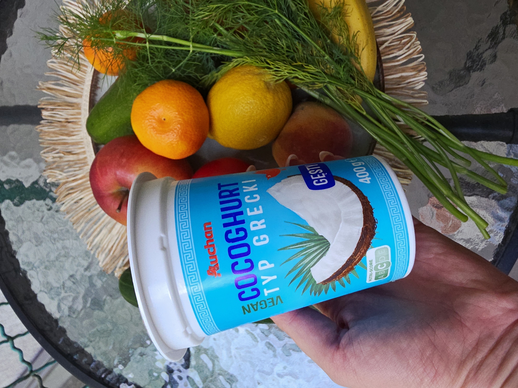 A close-up, high-angle shot captures a hand holding a container of coconut yogurt amidst a colorful assortment of fresh produce, all resting on a glass-topped outdoor table. The hand, visible in the lower right, firmly grasps the light blue and white yogurt container, presenting it towards the viewer.
The yogurt container's label prominently features the brand "Auchan" and the product name "COCOGURT TYP GRECKI," indicating a vegan, Greek-style coconut yogurt. Other text includes "VEGAN" and "GESTY," along with the weight "400G." A "NUTRI-SCORE A B C D E" is visible, with the letter 'A' highlighted in green, suggesting a high nutritional rating. The label also displays a stylized image of a coconut and palm leaves, bordered by a repeating Greek key pattern.
The produce is arranged on a circular, woven straw placemat, which sits on the textured glass surface of the table. Fruits visible include bright orange tangerines or oranges, yellow lemons, red apples, and reddish-yellow peaches. A green elongated vegetable, possibly a cucumber, is also present, and a yellow banana is partially visible in the upper right. Fresh green dill, with its feathery leaves, is scattered among the fruits, adding a vibrant green accent.
The bright, natural lighting indicates that the photo was taken during the daytime, likely outdoors on a patio or balcony, given the appearance of the glass table and its dark metal rim. The composition highlights the fresh ingredients and the vegan yogurt, suggesting a focus on healthy eating or a wholesome meal.