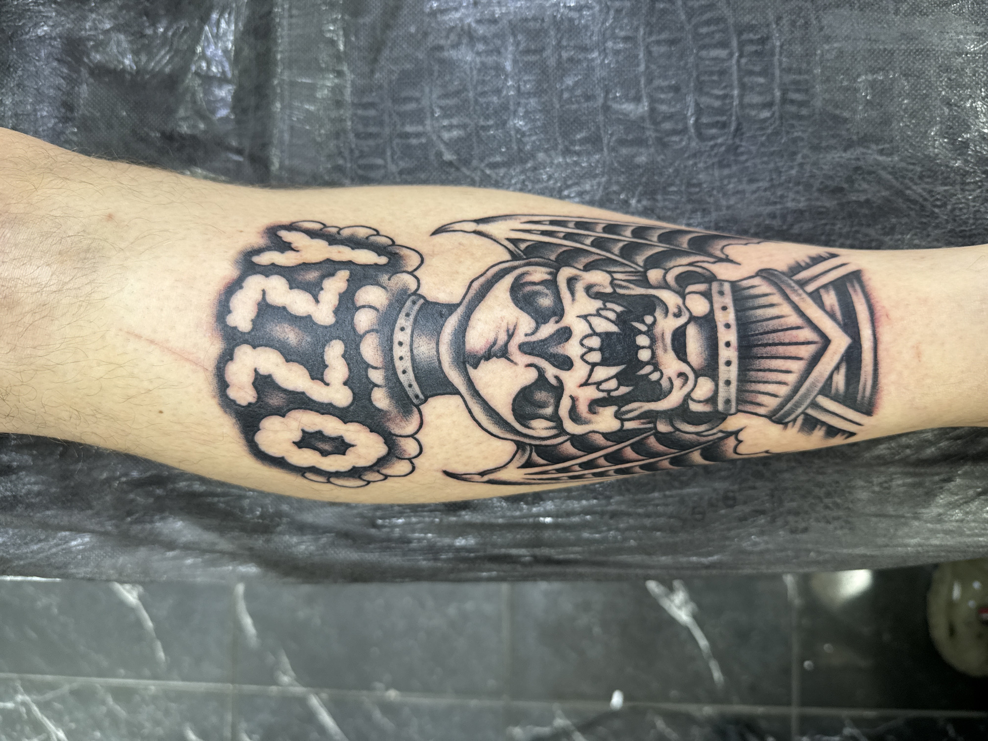 The image displays a close-up of a black and grey tattoo on a person's leg. The tattoo depicts a skull with bat-like wings, wearing a crown or headdress. Above the skull, the word "OZZY" is written in a cloud-like font. The background is dark and textured, suggesting a studio or backdrop, with some hints of a tiled wall and a grey fabric. The tattoo is the primary subject, rendered in a traditional American tattoo style. There is no identifiable location in Moscow, Russia, or any discernible action or interaction beyond the static presentation of the tattoo.