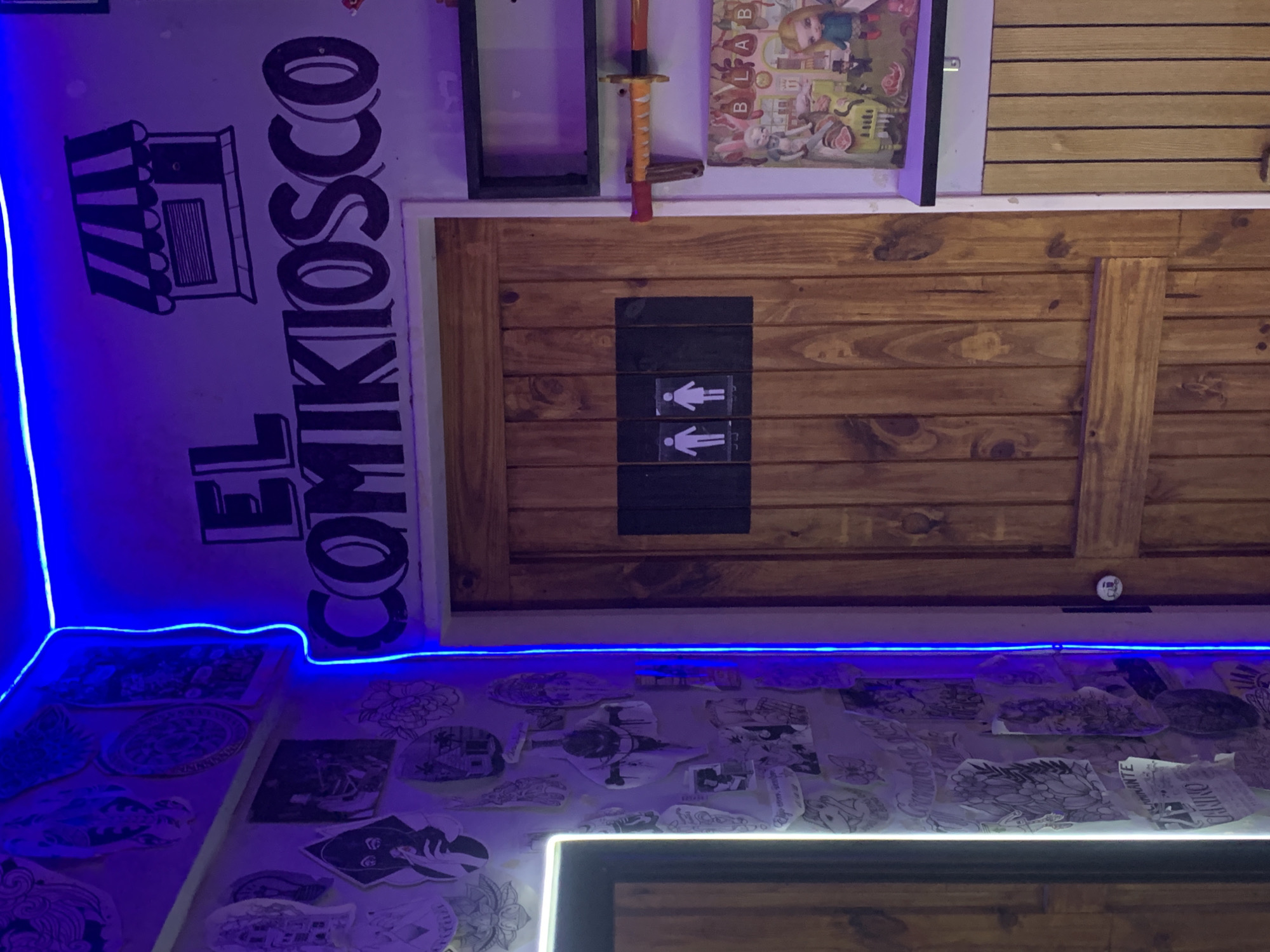 The image depicts the interior of El Comikiosco in Buenos Aires, Argentina. It appears to be a comic book store or related establishment based on the signage "EL COMIKIOSCO" in black lettering with an illustration of a kiosk to the left. The walls are adorned with various illustrations and sketches, possibly tattoo designs, attached to the surface. A blue LED strip outlines sections of the room. A wooden door with gender restroom symbols (male and female) is visible, along with other decorations, including a toy katana and a cartoon poster.