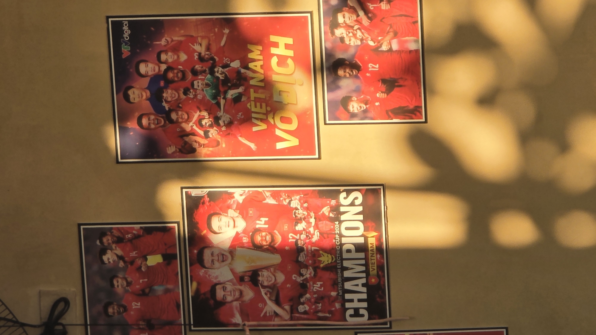 The image shows a collection of posters on a wall, presumably in Bắc Nha Trang, Vietnam. The posters feature the Vietnam national football team celebrating victories. One poster reads "VIỆT NAM VÔ ĐỊCH," meaning "Vietnam Champion." Another poster is for the "MITSUBISHI ELECTRIC CUP 2024 CHAMPIONS VIETNAM." Various players' faces are visible on the posters, along with their jersey numbers. The lighting in the photo suggests natural light and shadow patterns on the wall. A portion of what appears to be a black wire is seen in the bottom left corner. The wall itself is a light brown or beige color.