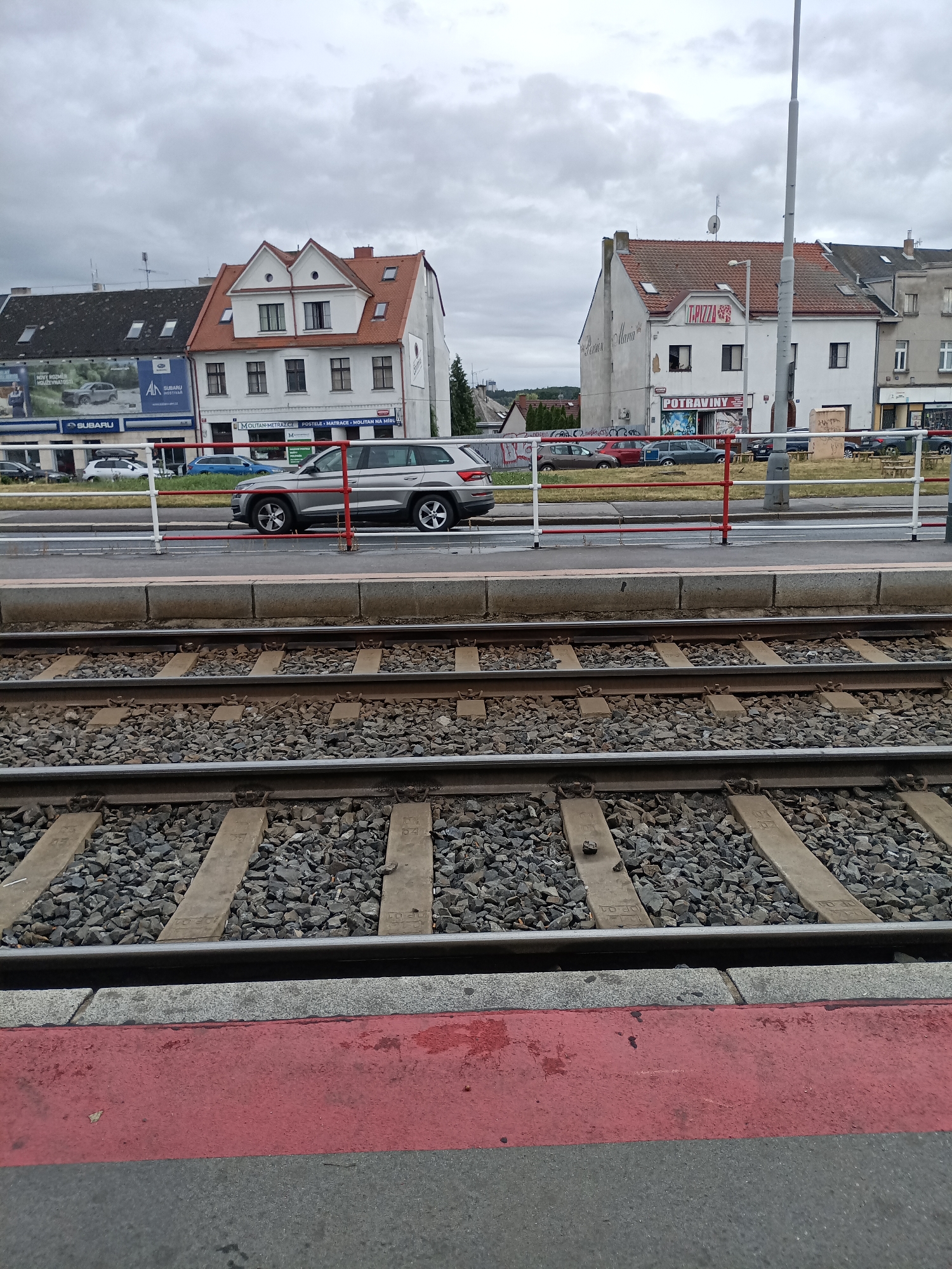 This is a medium shot, eye-level, outdoor photograph taken on a cloudy day. The foreground shows two parallel sets of train tracks with concrete sleepers and gravel ballast. A red stripe runs along the edge of the platform in the lower part of the frame. In the middle ground, a white and red striped metal barrier separates a road from the train tracks. Beyond the barrier, a silver SUV is parked on the right side of the road. The background features several buildings, some of which have red tile roofs. One building has signage that includes "POTRAVINY," indicating a grocery store, and another has "T. PIZZA" displayed. A large billboard for "SUBARU" is visible on the left side of the frame. The environment appears to be urban, possibly near a train station or public transportation hub in Prague, Czechia. There are no visible people or animals in the image.