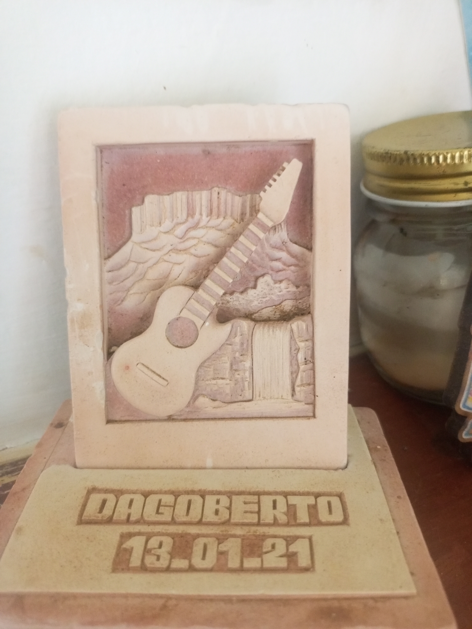 A close-up, eye-level, vertical shot shows a decorative item with a carved image of a guitar and a landscape featuring rock formations and a waterfall. The item is made of a light-colored material, possibly stone or plaster, and has text carved into its base. The text reads "DAGOBERTO" in the upper line and "13.01.21" in the lower line. To the right of the decorative item, a glass jar with a golden lid is partially visible. The background is a plain, light-colored wall. The scene appears to be indoors, on a wooden surface, suggesting it might be a souvenir or a decorative piece displayed on a shelf or table. There are no people visible in the image, and no specific details about the city of La Tagua, Venezuela are discernible from the image itself. The lighting is soft and diffused, with no harsh shadows.
