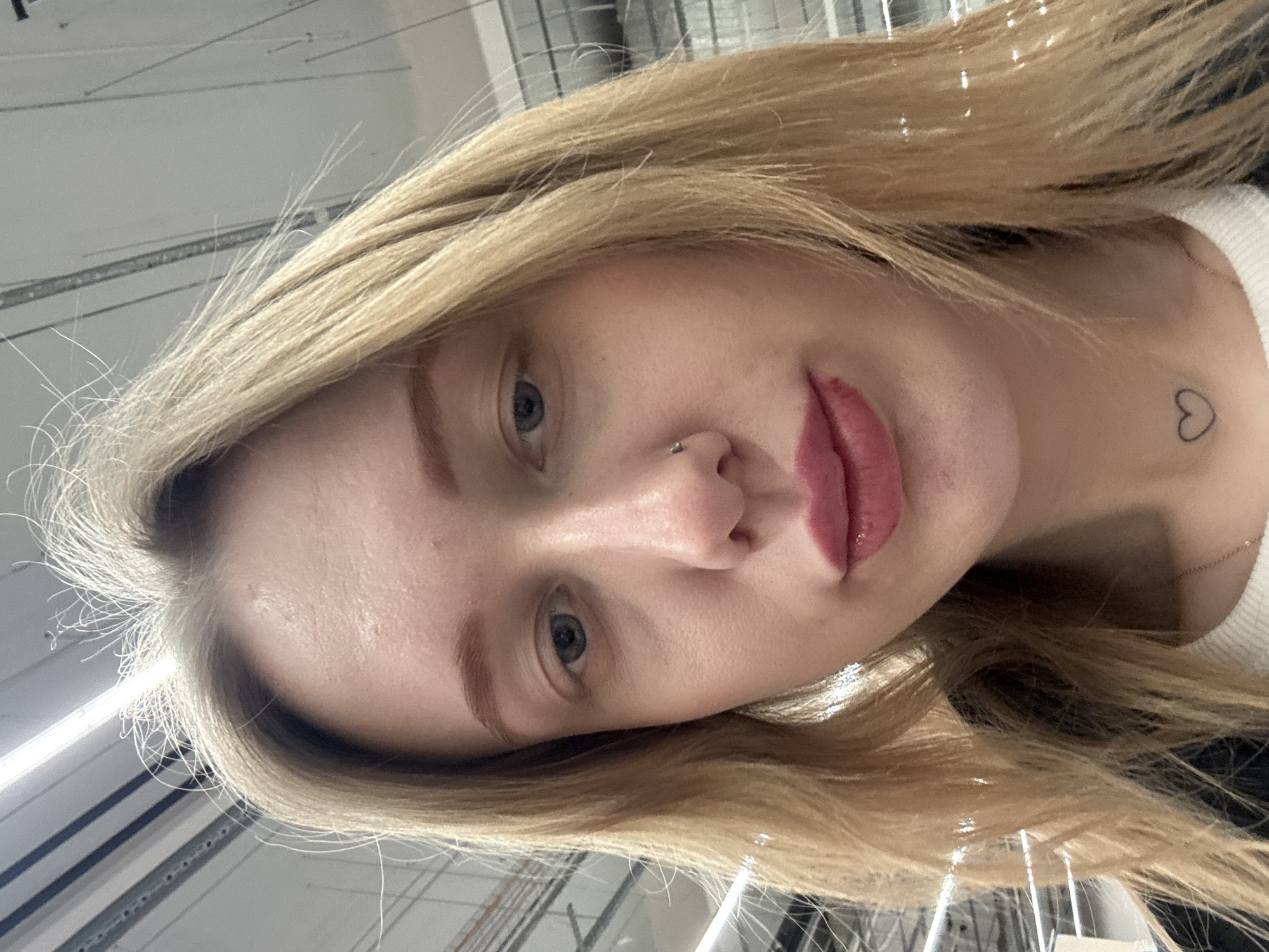 A close-up, low-angle portrait depicts a young adult female in an indoor commercial or industrial setting in Rostov-on-Don, Russia. The subject has long blonde hair, fair skin, and blue eyes, with reddish-brown eyebrows. A small silver stud piercing is present on her left nostril. Her lips are coated with a glossy pink substance, exhibiting a minor imperfection on the lower left. A small, black outline heart tattoo is visible on her neck, directly below her chin. She wears a white ribbed top underneath a dark outer garment, partially visible. The blurred background shows a ceiling with multiple parallel fluorescent light fixtures and exposed utility conduits, typical of a large retail space or warehouse.