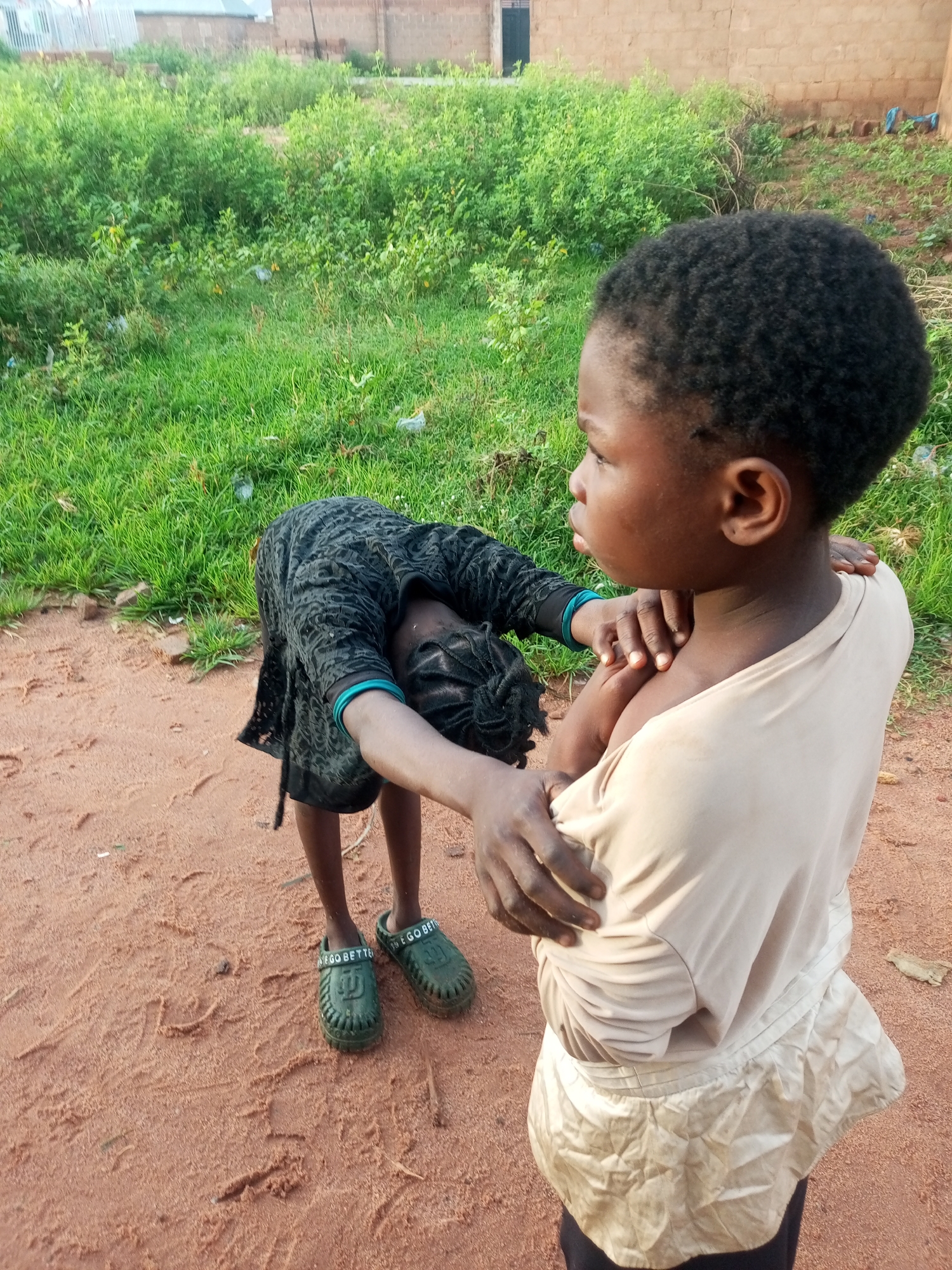 Two young girls are interacting outdoors on a dirt path in Tempure, Nigeria. The girl in the foreground, facing away from the camera, is wearing a light-colored t-shirt and a light-colored skirt. The second girl, in the background and bending over, is wearing a dark patterned dress with turquoise trim on the sleeves, and green clogs. The environment consists of a dirt path with footprints, dry grass, and some green vegetation in the background, as well as a brick building.