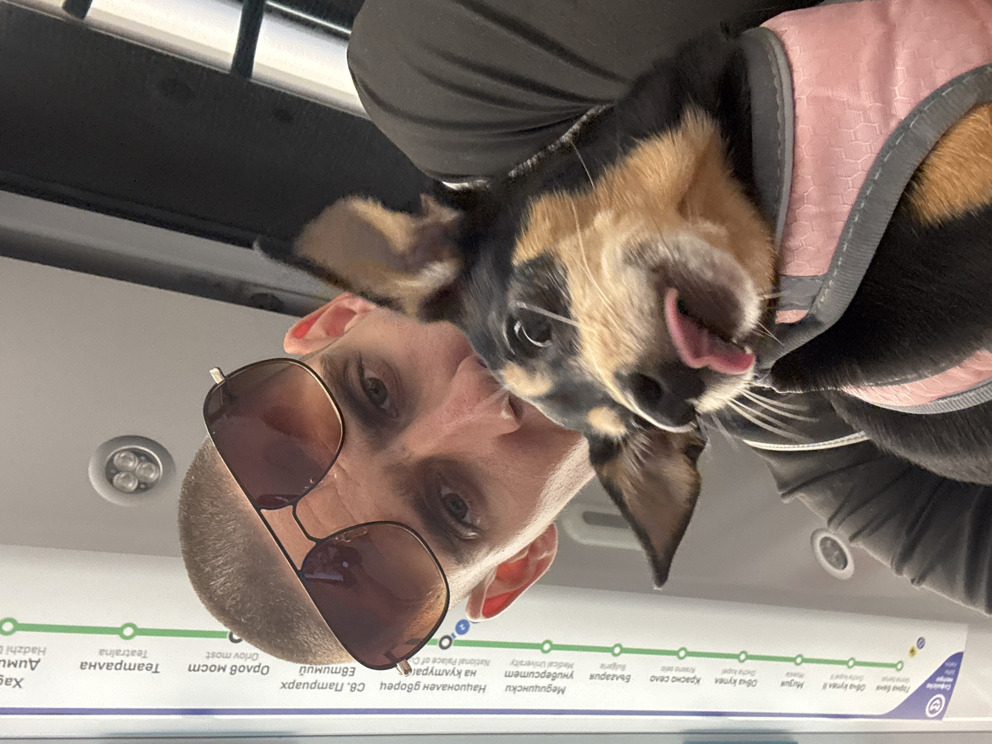 The image shows a close-up selfie of a man with sunglasses and a small dog in a pink harness. The man is looking at the camera, and the dog is sticking its tongue out. In the background, there is a map of the Sofia Metro system, indicating that the photo was taken on the Sofia Metro. The map shows station names like "Hadži Dimev", "Teatralna", "Orlov most", "Natsionalen Dvorets", "Meditsinski Universitet", and "Bulgaria". The man is wearing a black t-shirt or hoodie. The dog is mostly black and tan with floppy ears. The overall environment appears to be indoors, consistent with a metro train car.