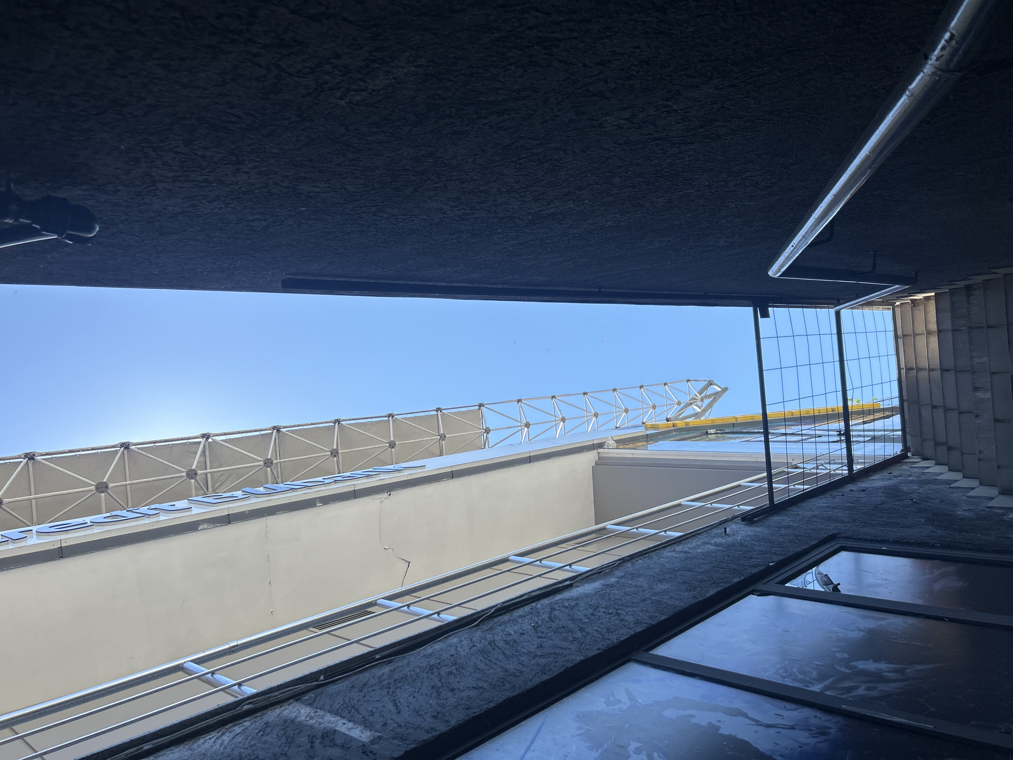 The image shows an outdoor view looking upwards towards a building and the sky. To the right, a dark, textured wall rises vertically, with a metal handrail running diagonally upwards along its surface. To the left, a portion of a building with a white facade and a balcony with metal railings is visible. Above the white facade, a light-colored triangular lattice structure extends across the top. The sky is clear and blue. In the foreground, a set of concrete stairs leads upwards between the dark wall and the building on the left, with a wire mesh fence along the edge of the stairs. The scene appears to be in an urban setting, possibly a narrow alleyway between buildings, in Burgas, Bulgaria. There are no people visible in the image.