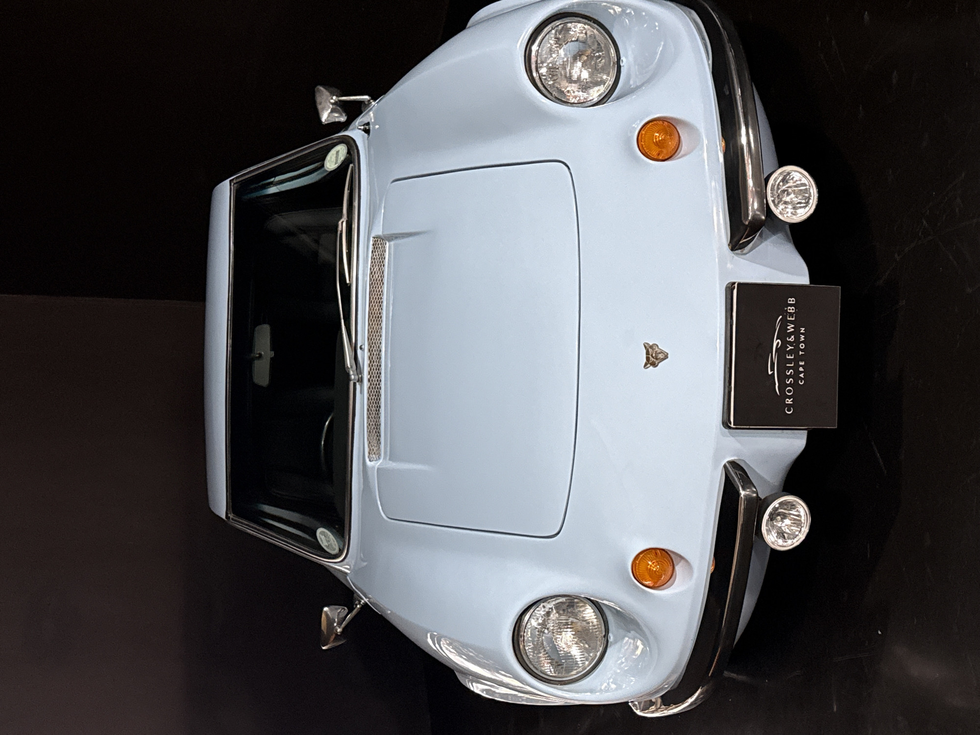 The image displays a light blue classic sports car from an overhead perspective, with its front facing forward, against a uniform dark background. This setting suggests an indoor environment such as a showroom or exhibition.
The vehicle is a two-seater convertible, featuring a glossy, pastel blue paint finish complemented by various chrome elements. Notable details include two large, round chrome-bezeled headlights and smaller, circular amber turn signal lights. A horizontal chrome grille is visible on the front bonnet, below which a small, stylized metallic emblem, possibly a feline, is affixed. Two auxiliary driving lights, also chrome and circular, are mounted on the front bumper.
The windshield and visible portions of the side windows appear dark, either tinted or reflecting the dark surroundings. A circular sticker is present on the driver's side of the windshield. A prominent black rectangular plaque with white lettering is positioned on the front right side of the car. The text clearly reads "CROSSLEY & WEBB" with "CAPE TOWN" below it, identifying the location as Cape Town, South Africa, and likely the car's vendor or restorer. The car is stationary, and no people are visible in the frame.