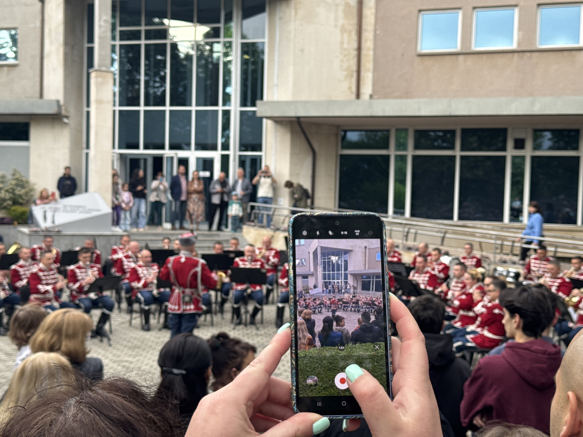 A person is holding a mobile phone in their hands, taking a video of a brass band performing outdoors. The band members are wearing matching red and white uniforms. They are seated in chairs arranged in rows on a paved area. Behind the band, a group of people stands on a raised platform in front of a building. The building has a modern architectural style with large glass windows. The sky is overcast. Based on the architectural style and uniform of the band, the image appears to be taken in Sofia, Bulgaria.