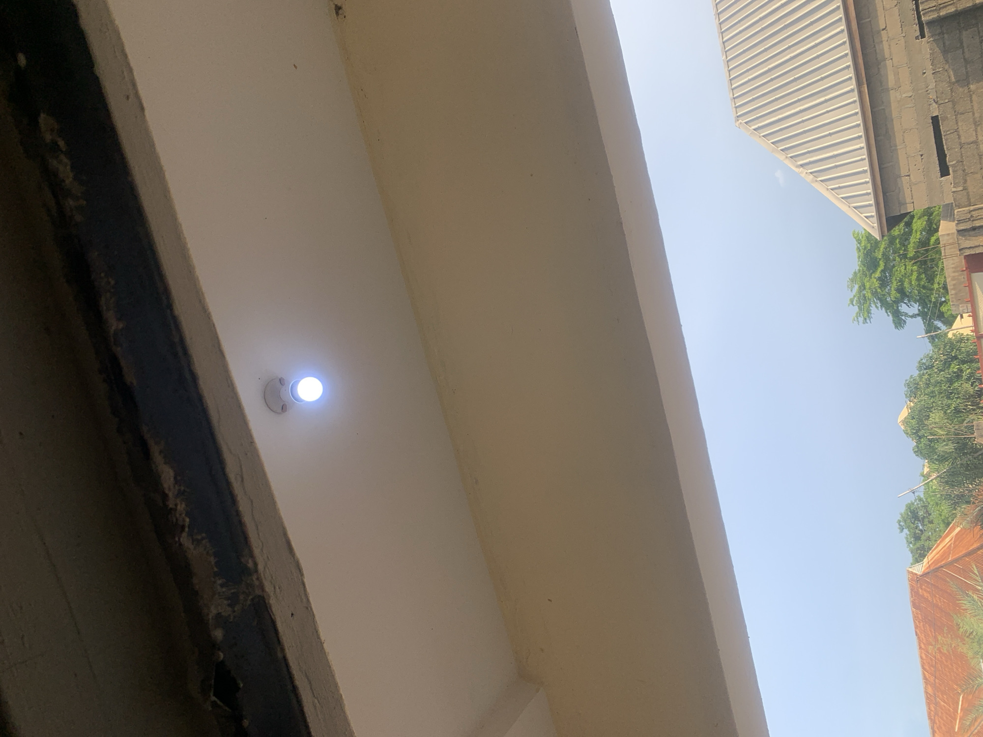 The image captures an exterior view under an overhang, looking upwards and outwards towards a hazy sky. A single, bright LED light bulb is illuminated and centrally positioned on the white ceiling of the overhang. Below the overhang, partially visible in the lower portion of the frame, are rooftops of buildings and green trees, suggesting an outdoor environment in a residential or semi-urban area. The sky appears overcast or hazy. The building materials visible include a corrugated metal roof and concrete blocks, characteristic of construction in regions like Jauro Yinu, Nigeria, where such materials are commonly used for housing and infrastructure.