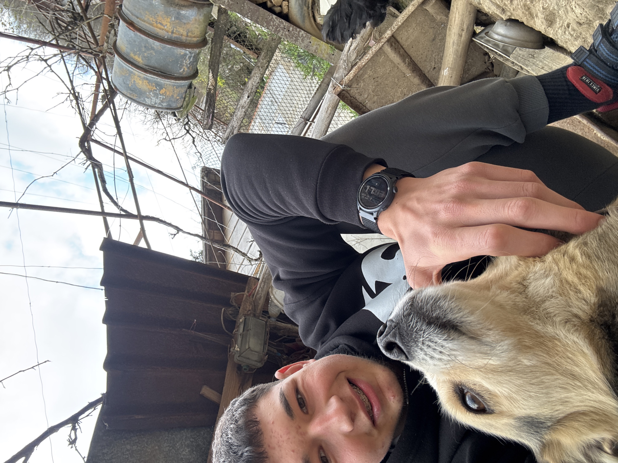 A selfie photograph taken outdoors shows a young male with braces, wearing a black hoodie, pants, and a black wristwatch on his left hand. He is smiling and positioned to the left of the frame, with a medium-sized dog nestled against his neck to the right. The dog has brown and tan fur. The time displayed on the wristwatch is 17:03.
The setting includes corrugated roofing, a metal drum-like object, wooden beams, and a wire mesh fence in the background, possibly within an agricultural or semi-rural area. The sock, visible above the shoe, reads "SINCE 1916". The location is presumably Zlatograd, Bulgaria.