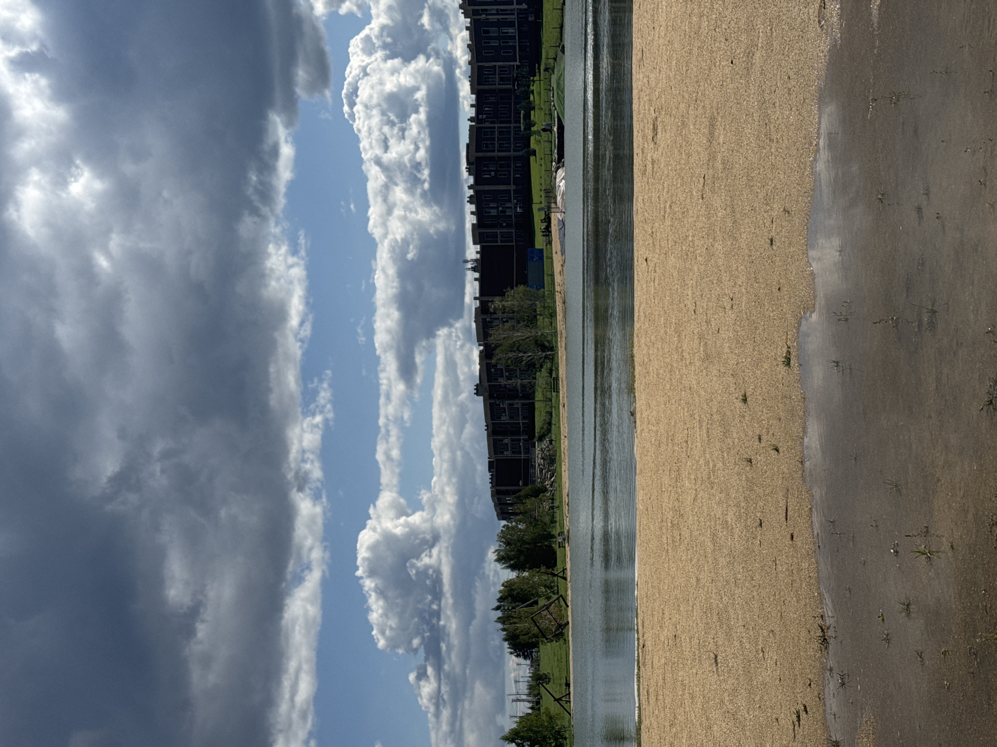 The image depicts a waterfront scene with a sandy beach in the foreground. Beyond the beach lies a body of water, and further back, a row of dark, multi-story buildings runs horizontally across the frame. Trees and some infrastructure, possibly part of a recreational area, are visible between the water and the buildings. The sky is filled with a mix of dark, heavy clouds and lighter, brighter clouds, suggesting a dynamic weather pattern. The sand appears to be a light tan color and is slightly wet in some areas near the water's edge. A puddle of water reflects the sky and the surrounding environment in the lower right portion of the sandy area. There are no discernible people or specific actions taking place in the visible portions of the image that would indicate a particular event or activity. The overall impression is of a somewhat overcast day at a lakeside or riverside developed area, with a focus on the natural elements of sky and water contrasted with the urban architecture in the background.