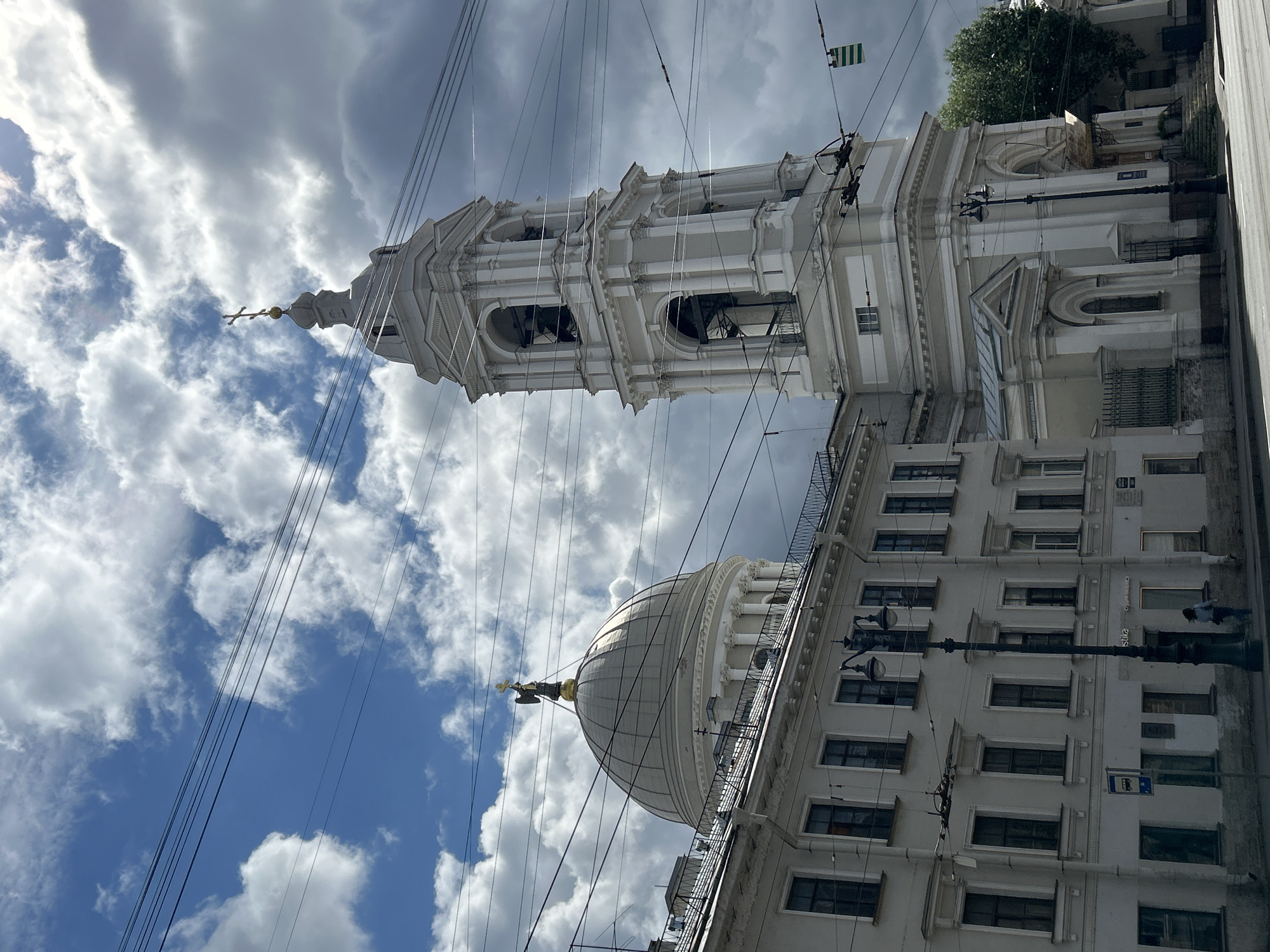 The image depicts a view of the St. Petersburg Stock Exchange building and its adjacent bell tower, known as the Rostral Columns, under a partly cloudy sky. The Stock Exchange building, a Neoclassical structure with a large dome topped by a statue of Neptune, is on the left. The bell tower, an ornate white structure with multiple levels and arched openings, is on the right, adorned with a golden cross. Several power lines crisscross the sky in the foreground, obscuring parts of the buildings and the sky. The lower portion of the image shows a street with a sidewalk and a few people walking. The architecture suggests a historical district within Saint Petersburg, Russia.