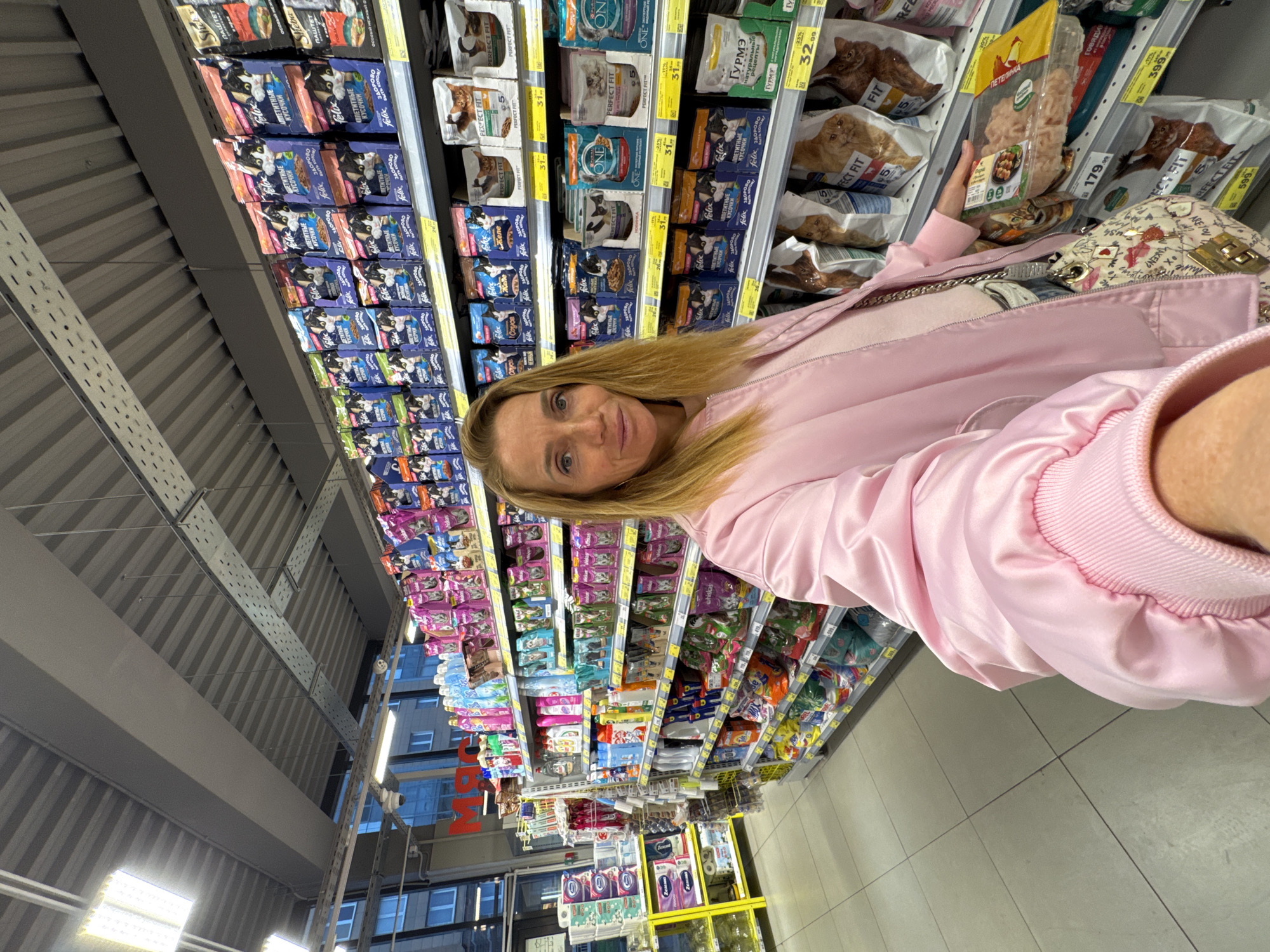 A woman is in a grocery store in Saint Petersburg, Russia. She is standing in an aisle with shelves full of pet food, mostly for cats. She is wearing a pink jacket and holding a package of pet food. The shelves are stocked with various brands and flavors of wet and dry cat food, including Felix, Perfect Fit, Sheba, and Gurme. Price tags are visible on the shelves. The store appears to be well-lit with a high ceiling. Through the exterior windows of the store, buildings in the city are visible.