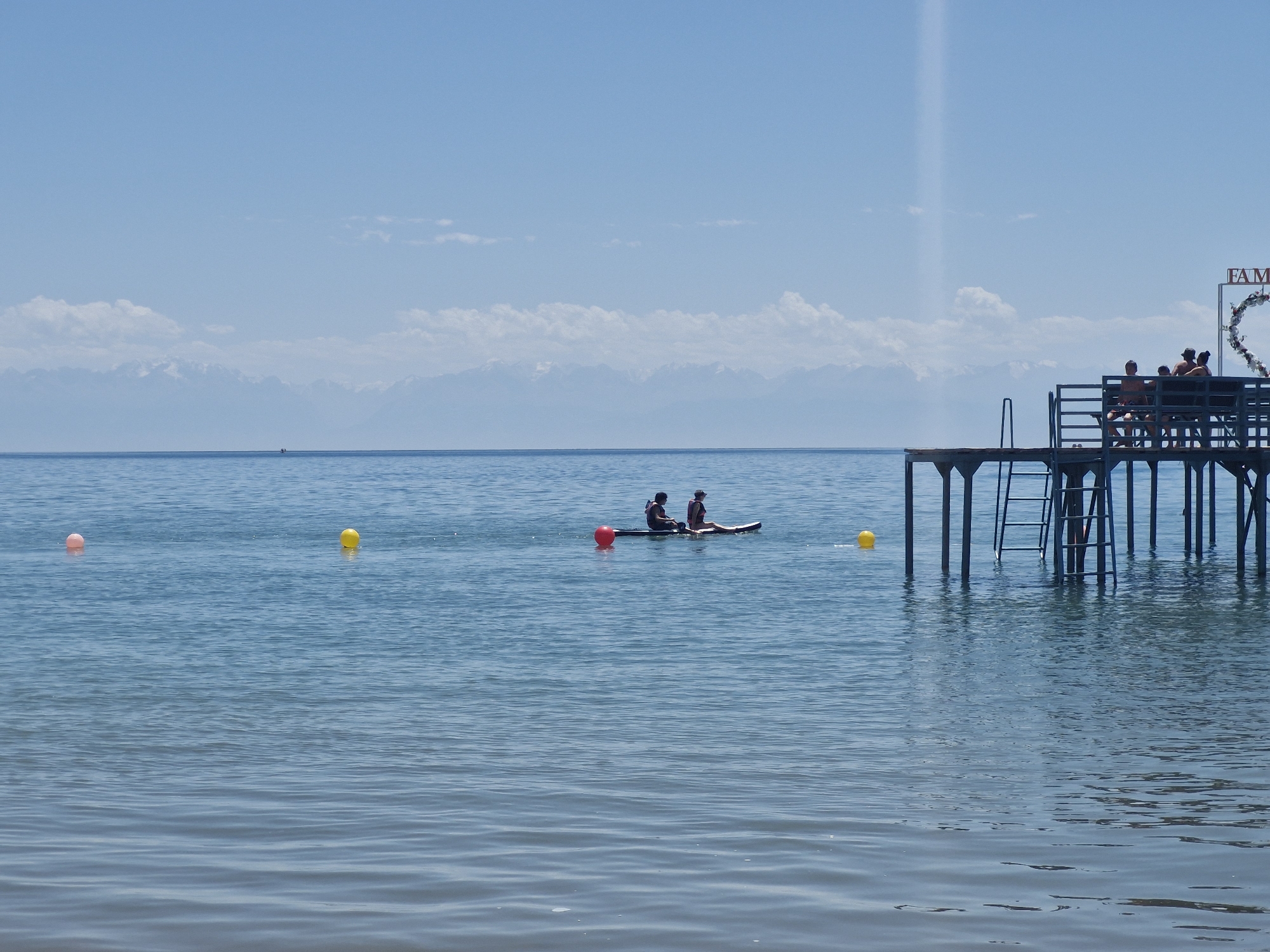 The image depicts a tranquil scene at Lake Issyk-Kul, near Bulan-Segettu, Kyrgyzstan. A wooden pier extends into the calm, blue water, where several people are gathered. On the pier, a group of individuals are seated on a bench, enjoying the view. To the left of the pier, two people are visible on a paddleboard in the distance, their forms silhouetted against the bright sky. The water is dotted with a yellow and a pink buoy. In the background, a range of snow-capped mountains forms a majestic horizon under a clear blue sky with scattered clouds. A decorative heart shape adorned with flowers is visible on the pier, alongside a sign that partially reads "FAM".