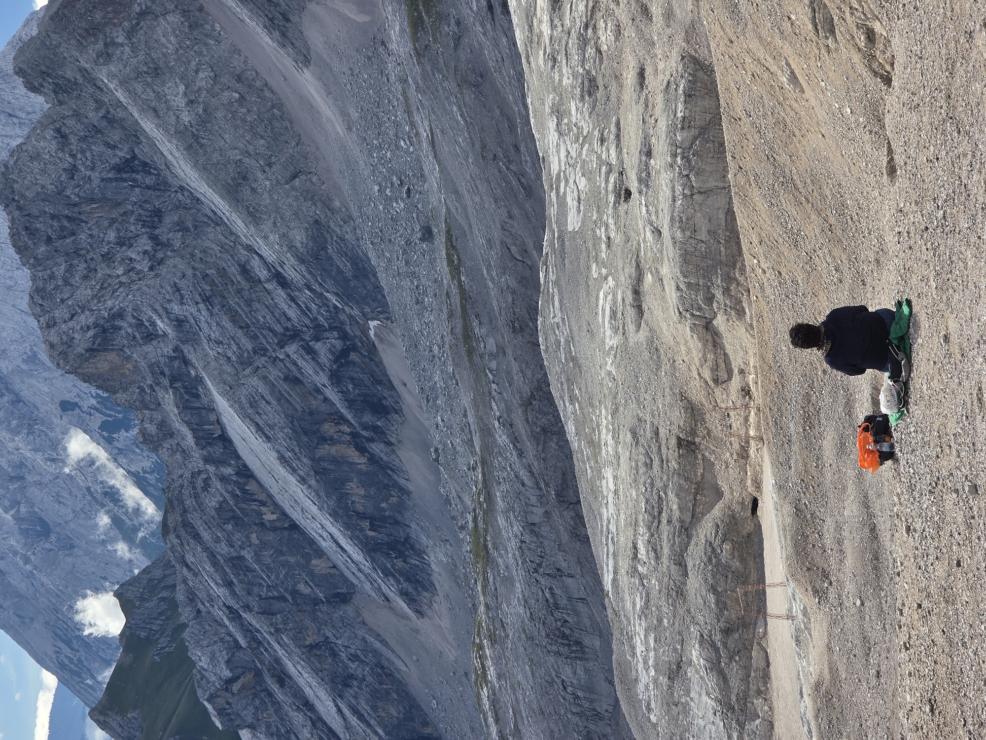 The image depicts a person sitting on the ground, facing away from the camera, with their back to a vast, rugged mountain landscape. The person is dressed in dark clothing and is seated on a green blanket with a backpack and an orange bag beside them. The environment is rocky and barren, characteristic of a high alpine region. In the background, large, grey, stratified mountains rise dramatically, with some patches of green vegetation visible on the lower slopes. Wisps of clouds are present in the sky and around the peaks. The scene suggests a moment of quiet contemplation or rest amidst the grandeur of nature. While the image does not explicitly show any urban elements of Garmisch-Partenkirchen, the mountainous terrain is consistent with the geography surrounding the city, which is nestled in the Bavarian Alps.