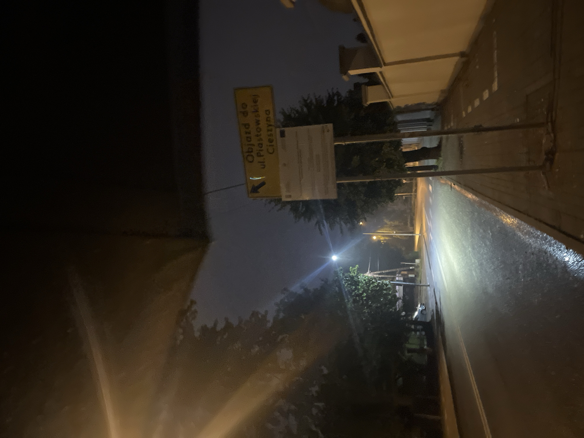 The image depicts a street scene at night, with overhead illumination casting long shadows and highlighting the wet pavement. Trees line the left side of the road, and a sidewalk is visible on the right. A street sign is mounted on a pole, displaying Polish text that indicates a detour. The sign reads "Objazd do ul. Piastowskiej Cieszyna," which translates to "Detour to Piastowska Street Cieszyn." A car is partially visible parked on the side of the road near the trees. The environment appears to be an urban or suburban area, possibly a residential street, given the presence of a building with a decorative wall on the right side of the image. The overall mood is subdued, characteristic of a dark and damp evening.