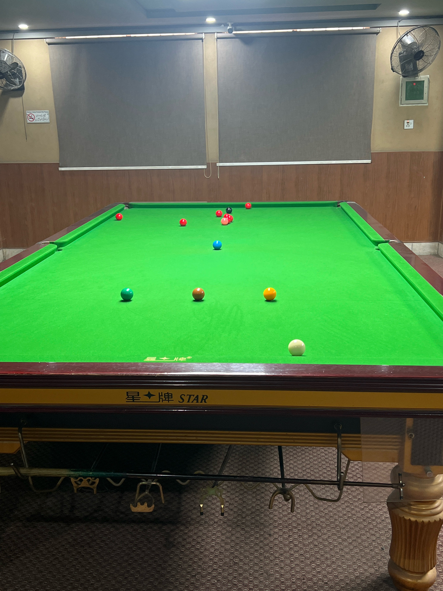 The image shows a snooker table in a room. The snooker table is green with various colored balls. The room appears to be indoors, with blinds on the windows and a fan mounted on the wall. There is also a sign on the wall that reads "No Smoking". The snooker table has "STAR" written on its side. This scene is located in Attock, Pakistan.