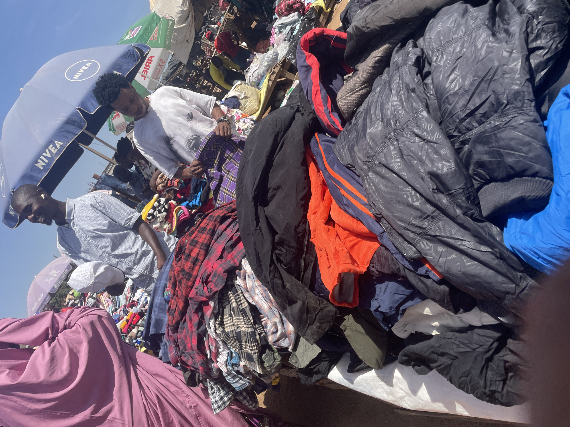 This image captures an outdoor market scene in Gura Top, Nigeria, bustling with activity under bright daylight. The main subjects are two men engaged in browsing through large piles of clothes. The man in the foreground, wearing a white short-sleeved shirt with dark patterns and a smartwatch, is leaning over, intently examining garments. Behind him, another man in a light blue patterned short-sleeved shirt, wearing wireless earbuds, is also looking through clothes.
The scene is dominated by vast heaps of clothing, primarily consisting of jackets, plaid shirts, and various other garments, suggesting a second-hand clothing market or a vendor selling bulk items. Notable items include dark padded jackets, some with subtle patterns, and colorful plaid shirts, as well as orange, blue, and black outerwear. The clothes are displayed on what appear to be tables or large mats.
The time of day is clearly daytime, with strong, direct sunlight creating pronounced shadows, indicative of midday. The sky is clear and bright blue. Overhead, a prominent blue umbrella provides shade, bearing the white text "NIVEA." In the background, another green umbrella is visible, showing mirrored text that reads "3ЯIА," which is "Ariel" reversed. The overall atmosphere suggests a busy, open-air shopping environment.