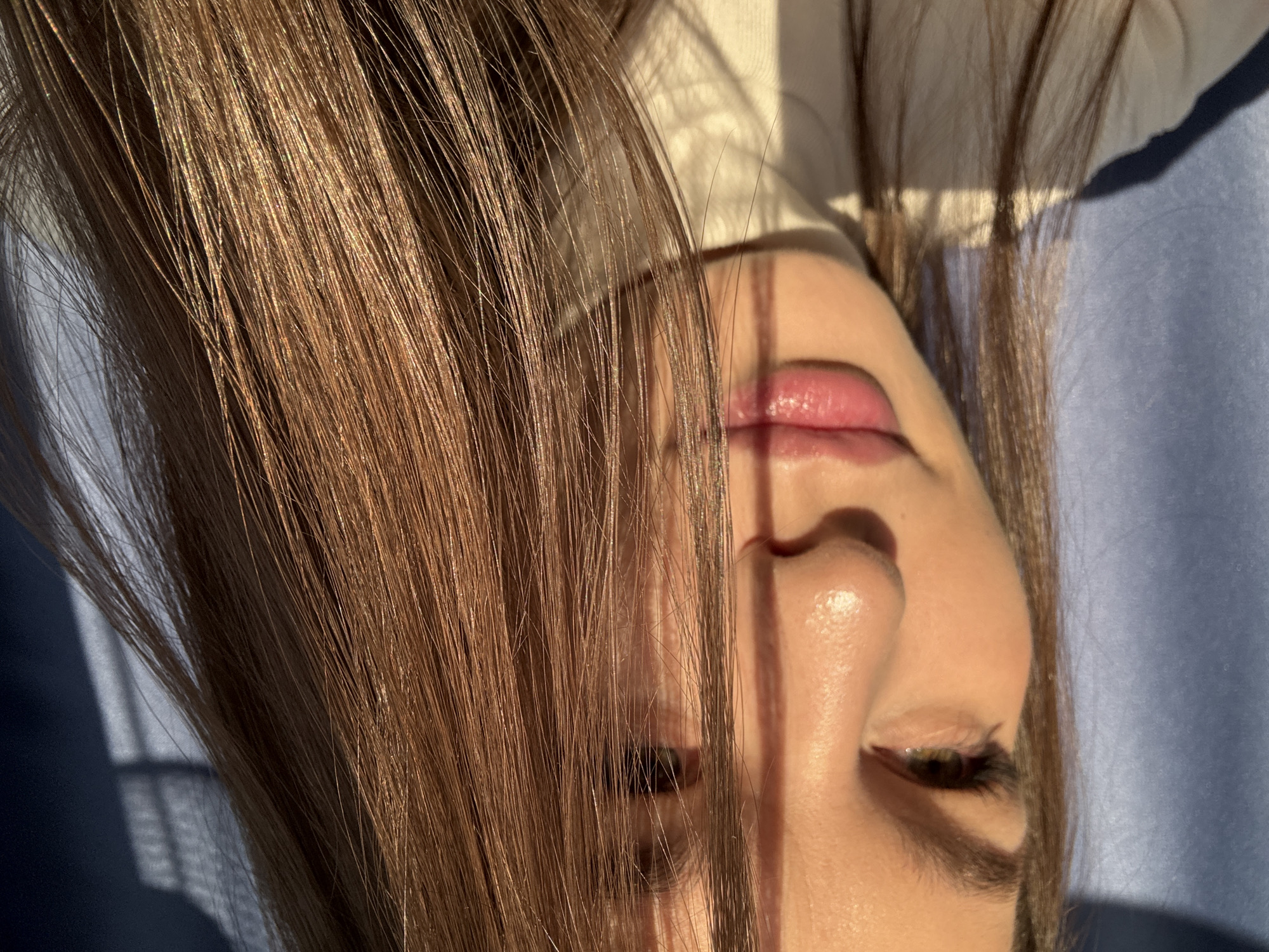 A close-up, slightly angled portrait captures the face of a fair-skinned individual, likely female, with dark brown hair partially obscuring the left side of their face. The hair is long and straight, catching the light. The person's right eye is visible, along with their nose and lips, which appear moisturized. Strong shadows and light cast by a window are visible across the face and on the blue surface below the person's head. The individual is lying on their back on what appears to be a blue surface. There is no immediately discernible information to identify the location as Chaikovsky, Russia, or any specific city.