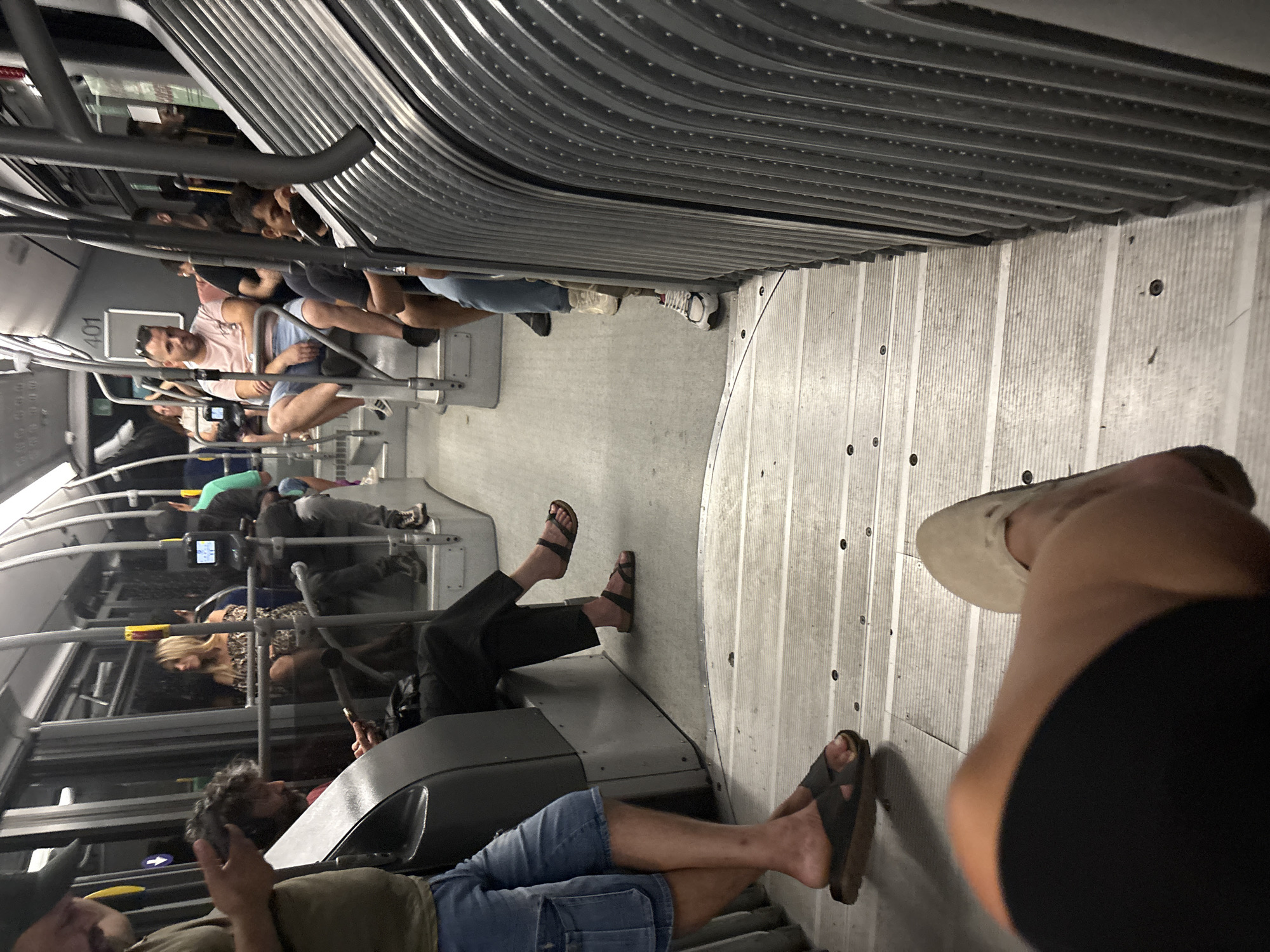 The image shows the interior of a public bus in Venice, Italy. Several passengers are seated and standing. In the foreground, a person's legs and feet are visible, with one leg bent and a foot wearing a light-colored sandal. To the left, a person wearing denim shorts and sandals is standing. Further back, several passengers are seated, including a woman with blonde hair wearing a patterned top, and a man in a pink t-shirt and blue shorts. The bus appears to be in motion, with the lights on and the interior illuminated. The seats are arranged along the sides of the bus, with a central aisle. The flooring is a light-colored, textured material. The bus displays the number "401" on an interior panel. The scene is a typical view of public transportation within the city.