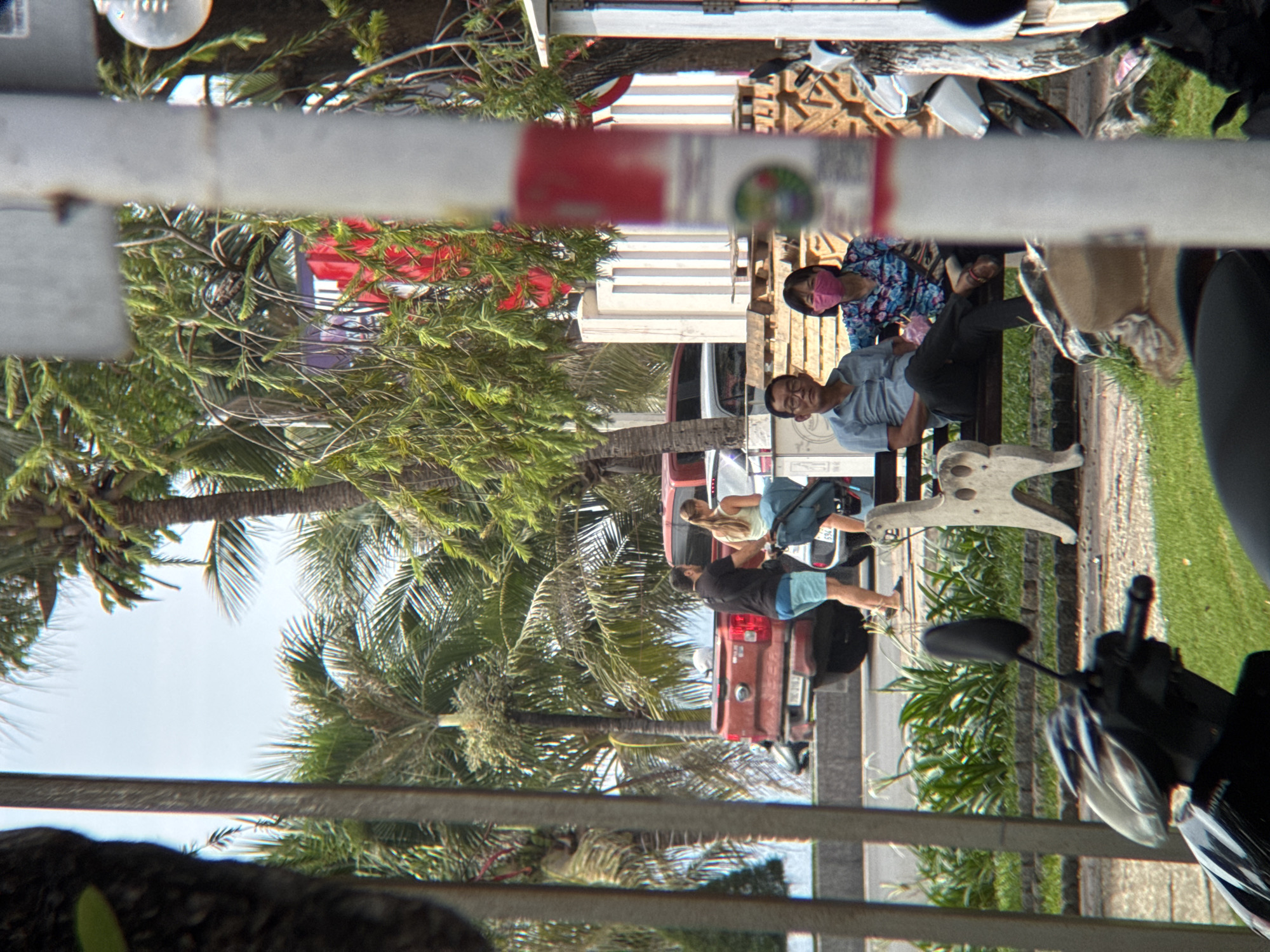 In Nha Trang, Vietnam, a man and a woman, both wearing face masks, sit on a dark bench in an outdoor setting. The man, on the left, wears a light blue collared shirt and dark pants. The woman, on the right, wears a brightly patterned shirt. Behind them, a white concrete statue with animal-like features is visible. In the background, a red pickup truck is parked. Several palm trees are scattered throughout the scene, indicating a tropical environment. A motorcycle is partially visible in the foreground on the left. A white pole with a red stripe is positioned in the center right of the frame.